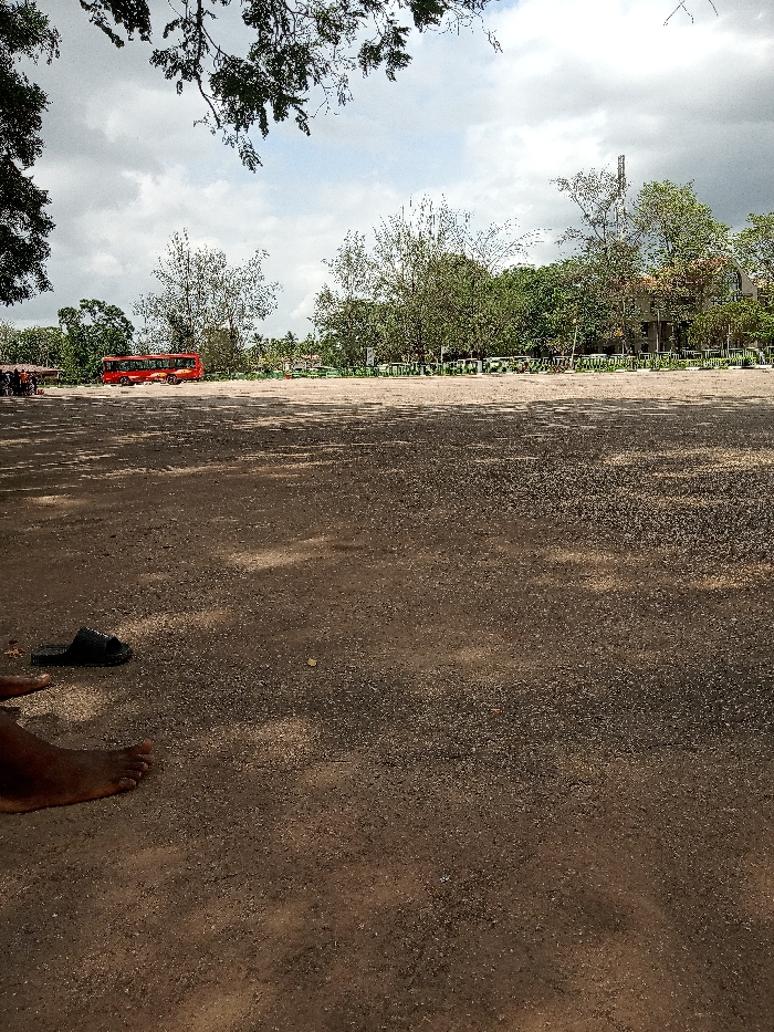 The image shows a large, empty dirt lot. The lot is located at coordinates 6.676 latitude, 3.165 longitude, which is in the city of Ota, Nigeria. A red bus is parked in the distance, and a small, sparsely populated green area is visible in the background. There are a few trees surrounding the lot. A pair of bare feet and one black sandal are visible in the foreground. The ground is dry and dusty. The sky is partially cloudy. The image was taken during the daytime.