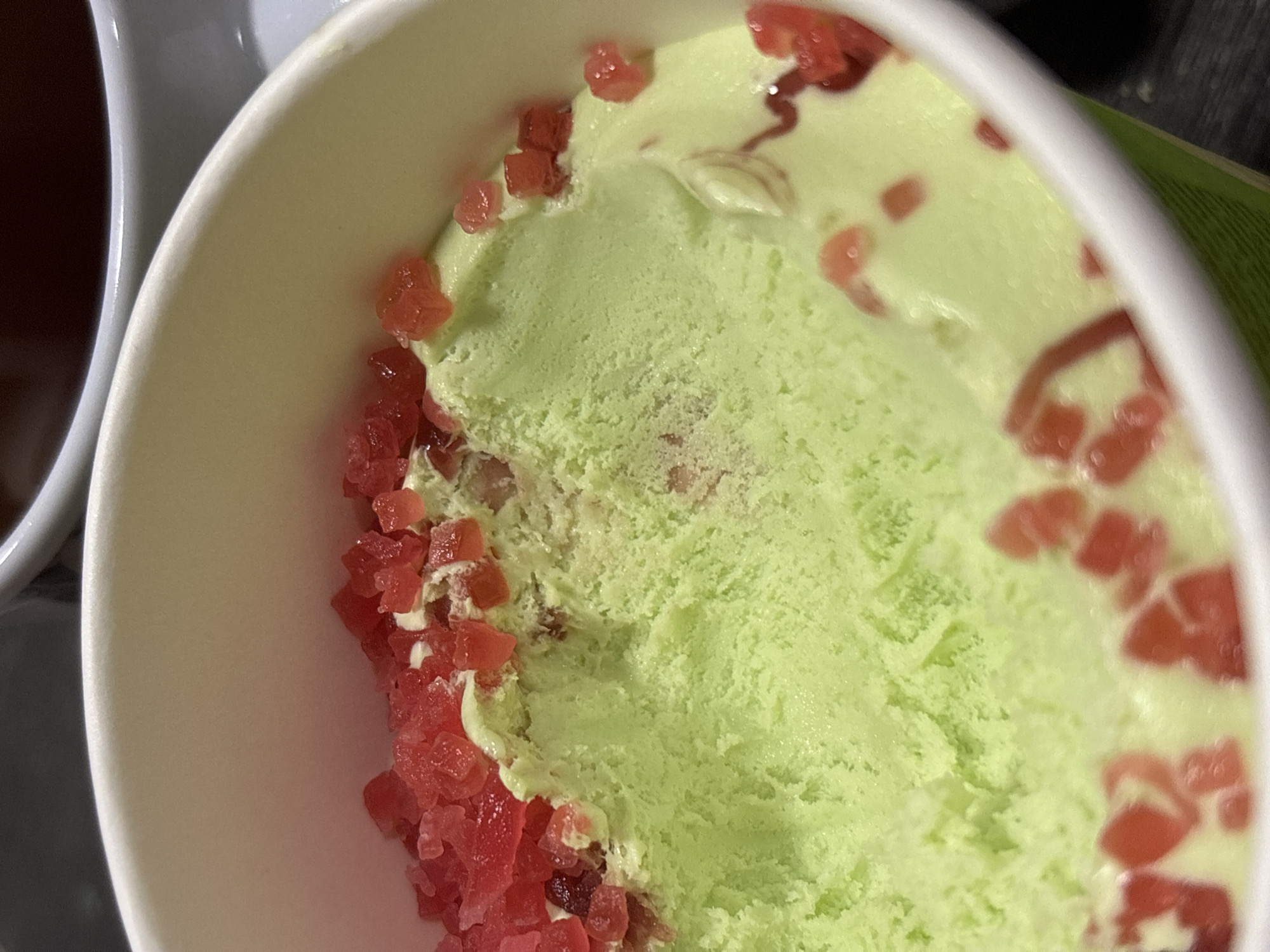 A close-up shot depicts a white paper cup filled with pale green ice cream, topped with small red cubes. Part of another white cup containing a dark liquid is visible to the left of the ice cream. Based on visual analysis, the location cannot be determined, nor is there any indication of it being Perm, Russia. People, interactions and specific environmental details are not discernable.