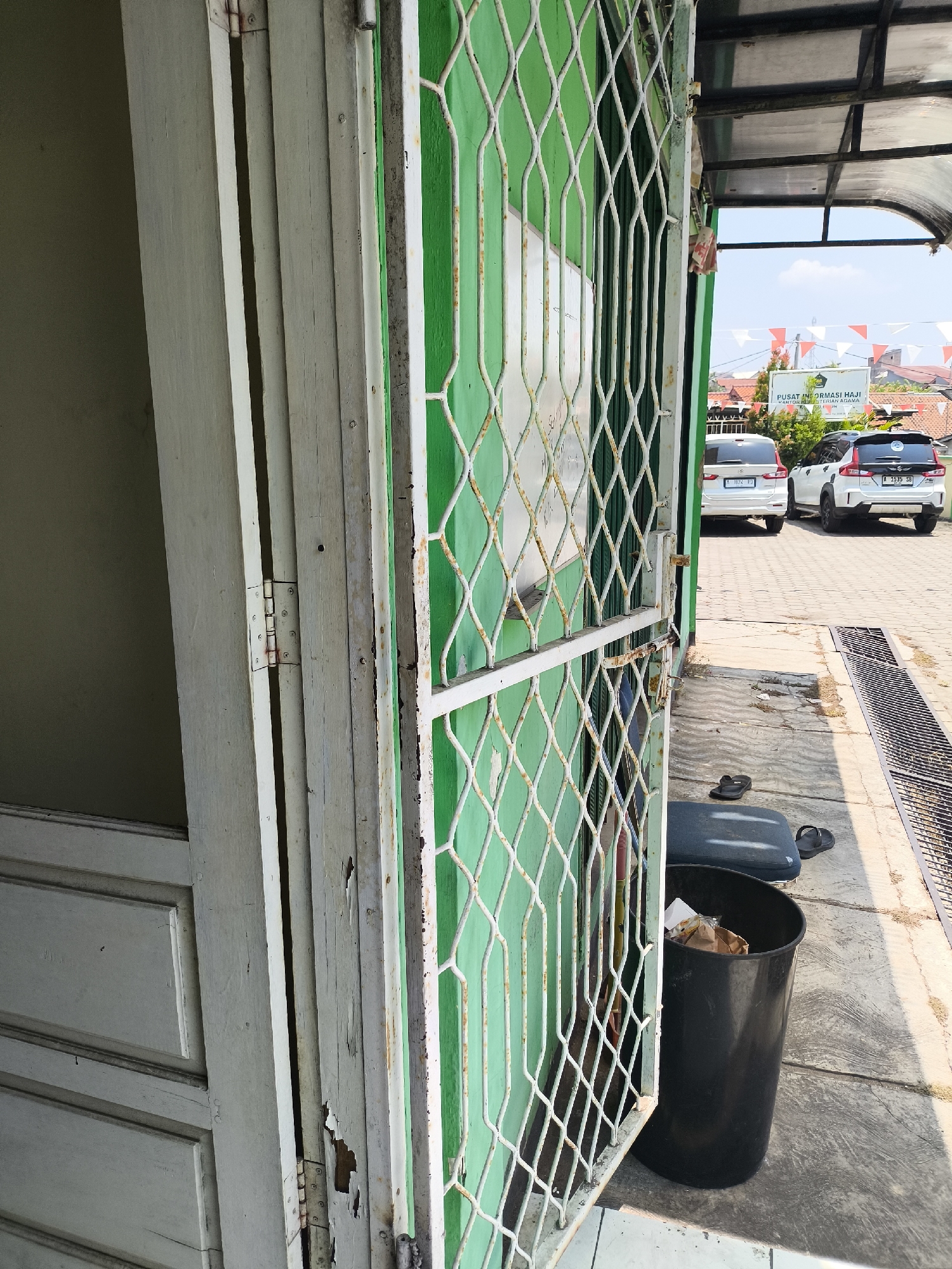 The image shows the exterior of a building with a green painted wall and a white metal gate. The gate is made of diamond-shaped mesh and appears to be slightly rusted. To the left of the gate is a wooden door that is partially open. The door is painted white and shows signs of wear and peeling paint. In front of the gate, there is a black trash can filled with refuse, and a dark blue cushion with a pair of black flip-flops resting on it. Behind the trash can and cushion, a drainage grate runs along the ground. In the background, two white cars are parked on a paved surface, and a building with a sign that reads "PUSAT INFORMASI HAJI" (Hajj Information Center) is visible. The sky is clear and blue, indicating it is daytime. The overall scene suggests an outdoor area in front of a public building. The location is identified as Cilegon, Indonesia.