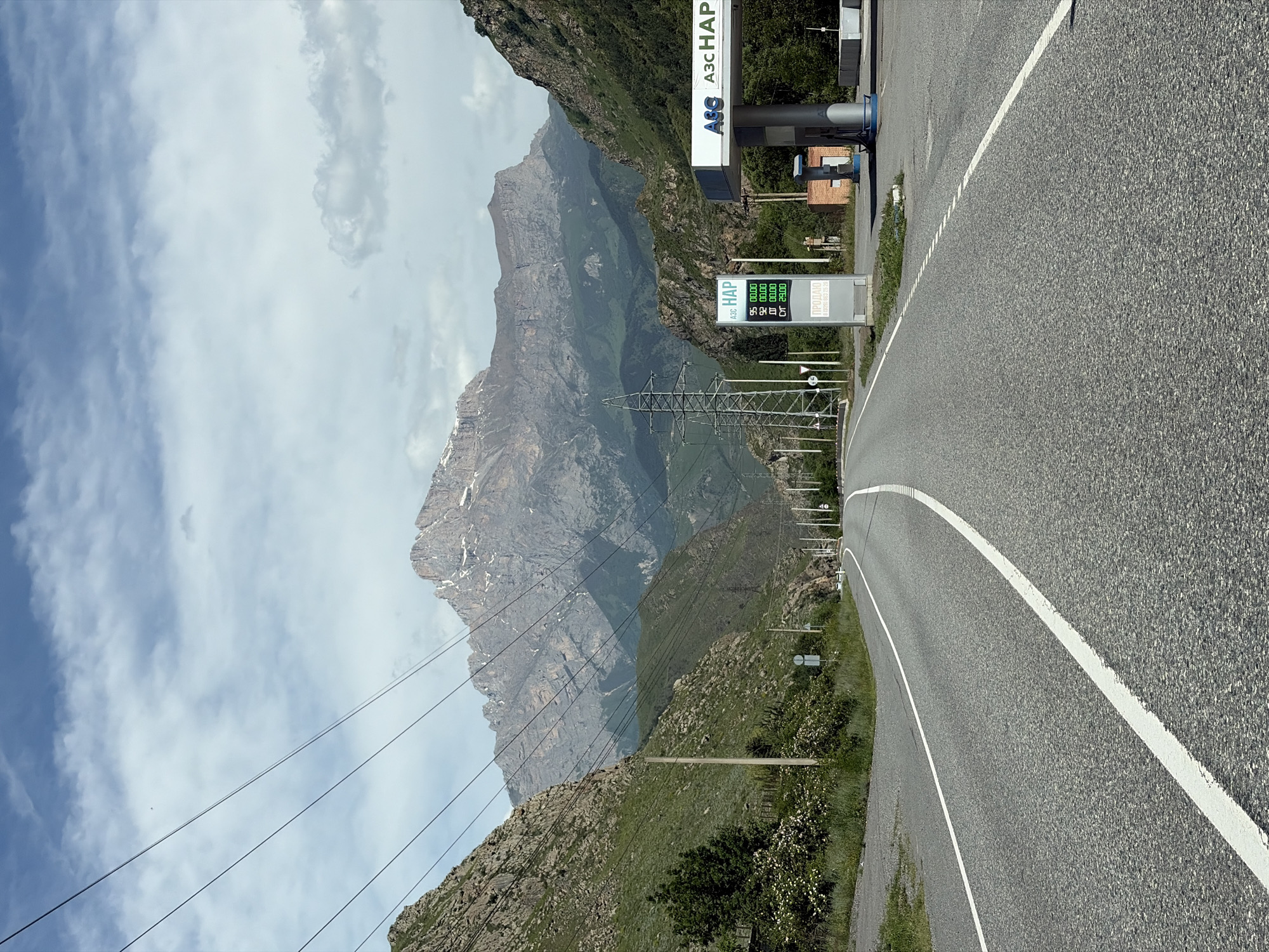 The image depicts a scenic road leading towards a towering mountain range under a cloudy sky. The road, a two-lane asphalt surface with white markings, curves to the right as it enters a valley. On the right side of the road, a gas station with a prominent blue and white sign reading "АЗС НАP" and digital price displays for fuel is visible. A white fuel dispenser is present. Power lines stretch across the sky, supported by tall metal towers. The mountains flanking the road are covered in lush green vegetation and rocky outcrops. The overall scene is one of a natural landscape with man-made infrastructure present. The location is the city of Mizur, Russia.