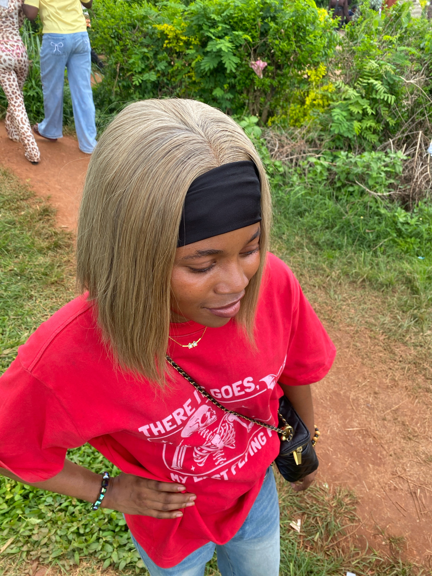 The image depicts an outdoor scene, likely on a dirt path adjacent to a grassy area with dense green foliage. The primary subject is a young Black woman, viewed from a high angle, looking downwards and slightly to her right. She has light brown hair, styled in a bob, and is wearing a red t-shirt with white text and graphics, possibly a skeleton motif. She is also wearing blue jeans, and a black quilted handbag with a gold chain strap is slung across her shoulder. On her left wrist, she wears a beaded bracelet.
In the background, to the left of the frame, two other individuals are partially visible. One person wears a yellow shirt and light blue jeans with a white bow detail on the back pocket. Another individual to their left wears a leopard-print outfit. The background suggests a natural, possibly rural or semi-urban environment with abundant vegetation and a dirt pathway. The image captures a moment outdoors, suggesting a casual setting. The presence of people in light clothing and the lush greenery indicate a warm climate. The scene is set in the city of Ado Ekiti, Nigeria.
