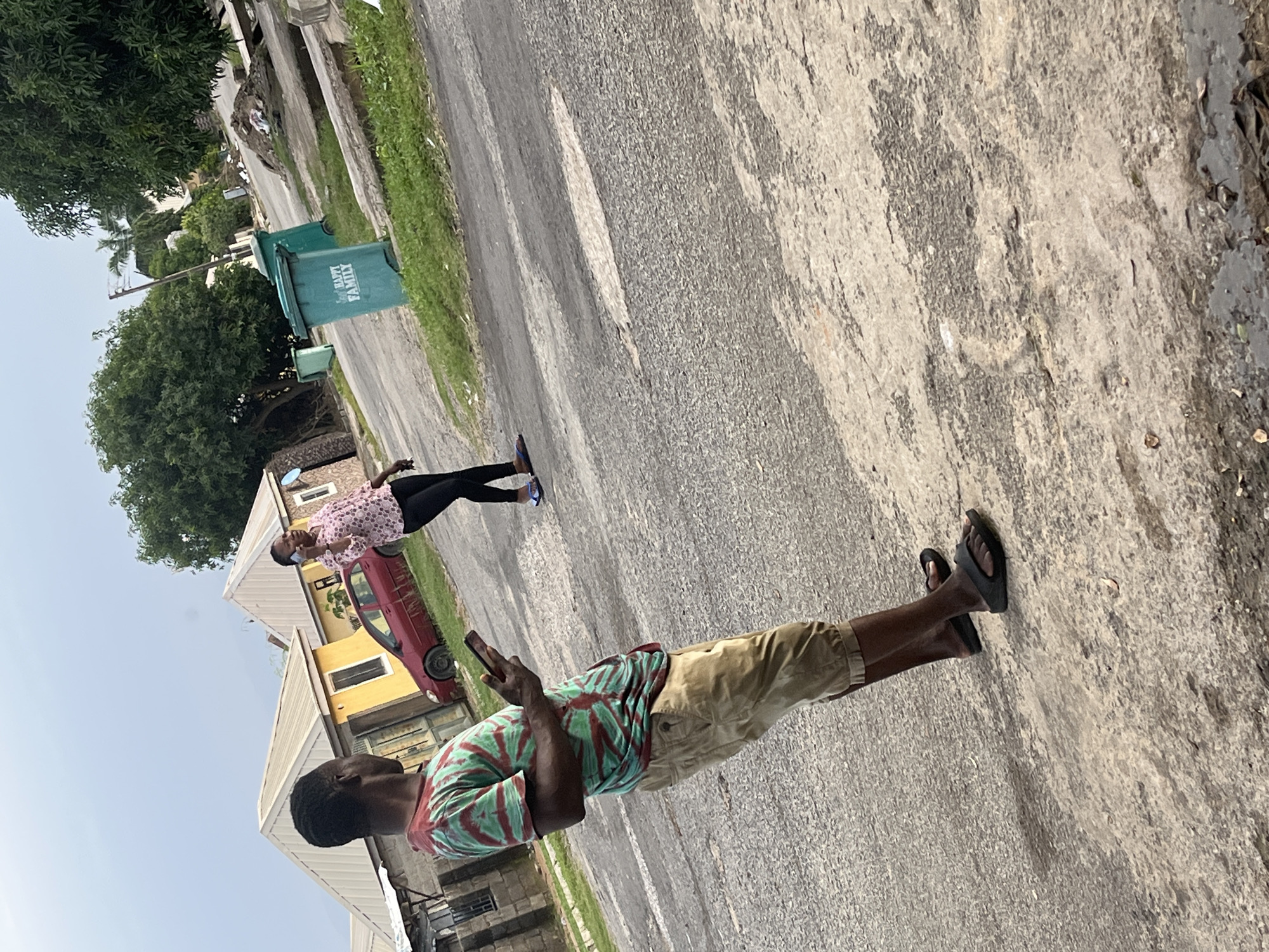In a residential area of Abuja, Nigeria, two individuals are present on an asphalt road. A man in the foreground, wearing a tie-dye t-shirt and khaki shorts, is holding a smartphone and looking down at it. A woman in the background, dressed in a patterned pink shirt and black pants, is walking away from the camera, speaking on her phone. Two green trash bins, one with "HAPPY FAMILY" written on it, are visible to the right of the road, near a pile of debris. Houses with sloped roofs and a red car are situated behind the man. The environment is overcast, suggesting it may be an early morning or late afternoon.