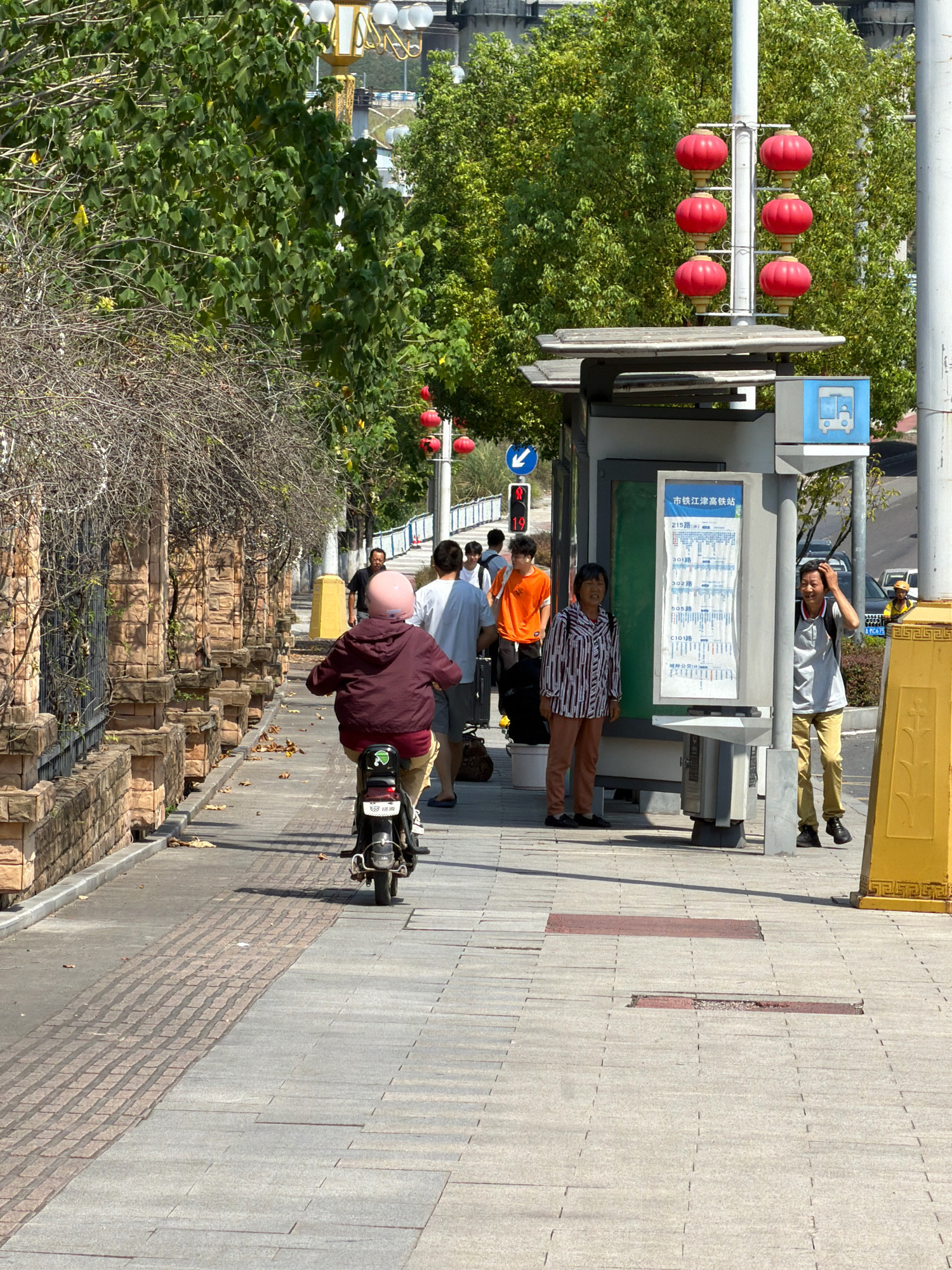 The image captures a street scene in Jiangjin, China. On the left, a stone wall with decorative pillars lines a sidewalk. A person on a scooter, wearing a burgundy jacket and a pink helmet, rides away from the camera. In the background, trees with lush green foliage line the street. To the right, a bus stop shelter stands with a route map displayed on a pole. Several people are gathered at the bus stop, including a woman in a striped shirt and brown pants, and a man wearing a gray shirt and khaki pants. A few other individuals are visible in the distance. The sidewalk is paved with gray tiles, with some sections featuring a reddish-brown brick pattern. The weather appears to be sunny, casting shadows on the ground. Red lanterns are suspended on a pole near the bus stop. A yellow decorative post stands on the right side of the image.