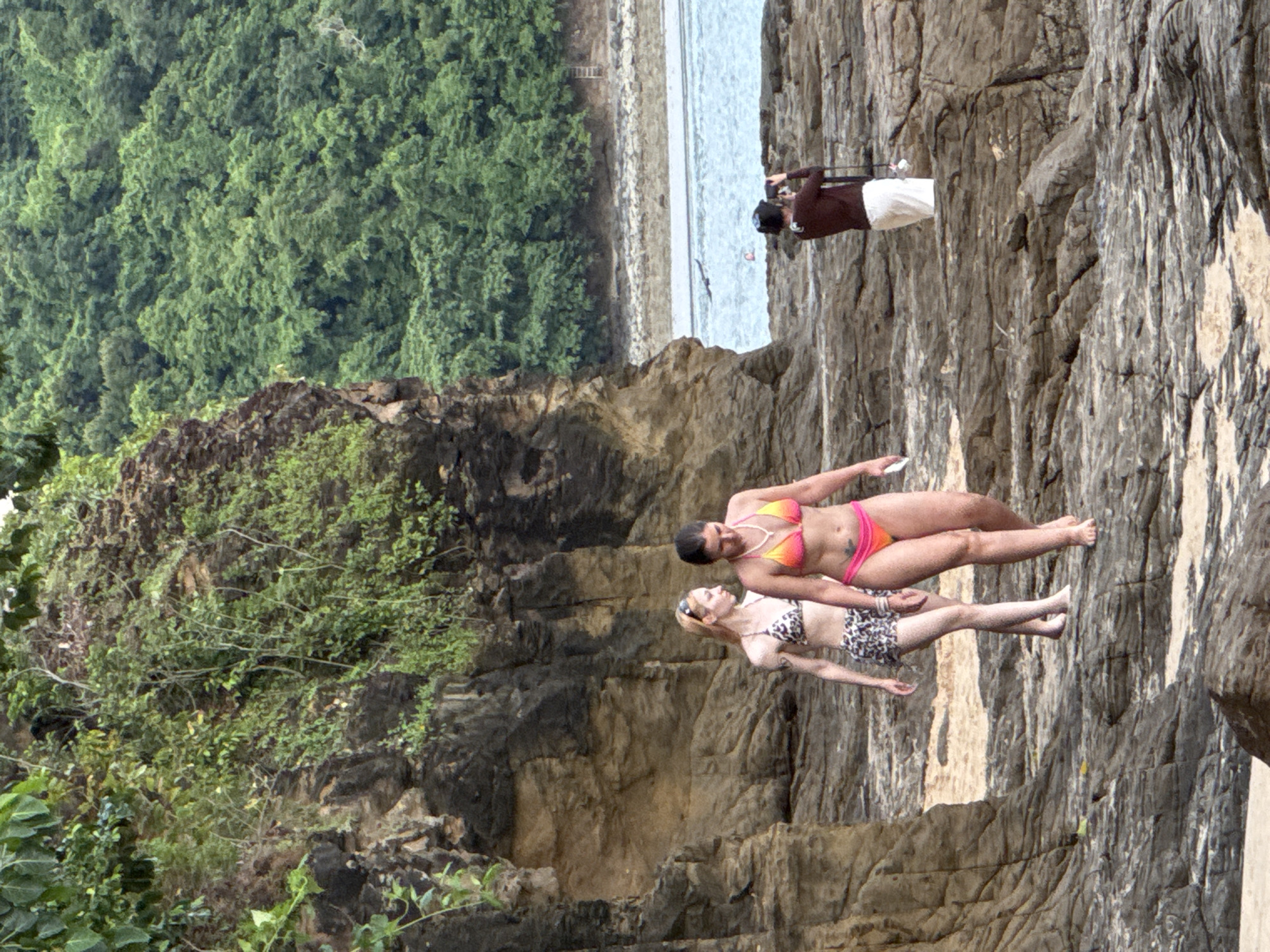 Two women in bikinis are walking on a rocky shore. The woman on the left has blonde hair and is wearing a black and white patterned bikini. The woman on the right has dark hair and is wearing a pink and orange bikini. Behind them, a person wearing a brown top and white pants is taking a picture with a camera. The background consists of a rocky cliff face with green foliage and a lush green forest. The ocean is visible in the distance. There is no information in this image to suggest it is located in the city of Wichit, Thailand.