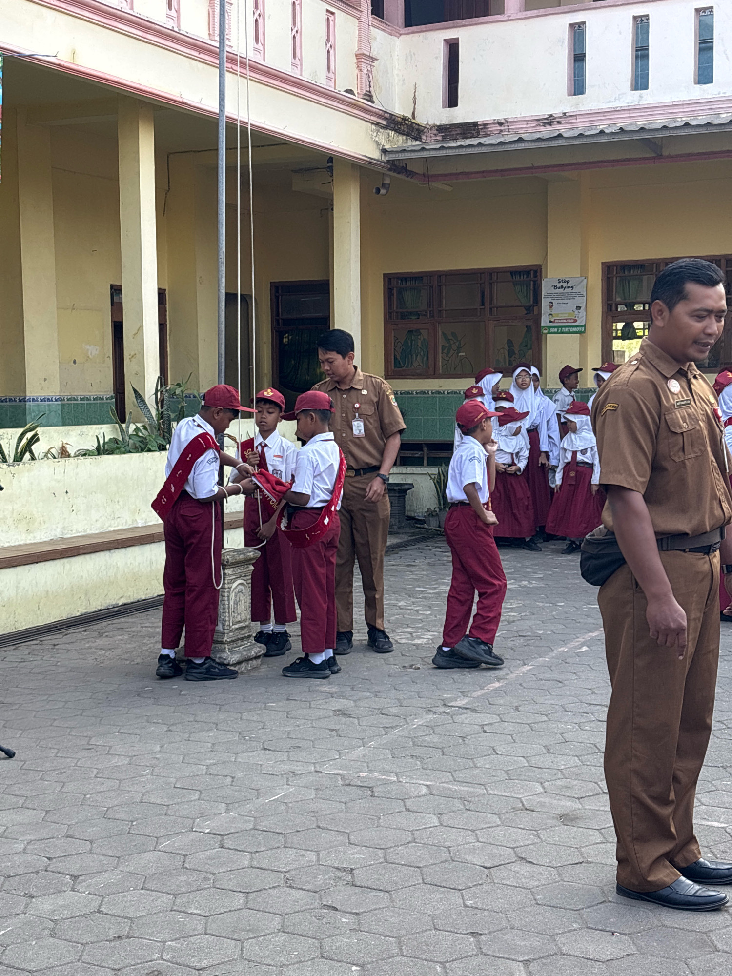 The image depicts an outdoor scene at a school in Tirtomoyo, Indonesia, likely during a flag-raising ceremony or assembly. In the foreground, a man in a brown uniform stands to the right, partially obscuring the view of several school children. The children, dressed in white shirts, red hats, and red pants or skirts, are gathered around a flag pole. Some of them appear to be holding or preparing a flag. A teacher, also in a brown uniform, stands behind the children, observing them. In the background, a school building with yellow walls and pink accents is visible. A poster with the text "Stop 'Bullying'" is displayed on the building. The ground is paved with hexagonal stones. The overall atmosphere suggests an organized school event.