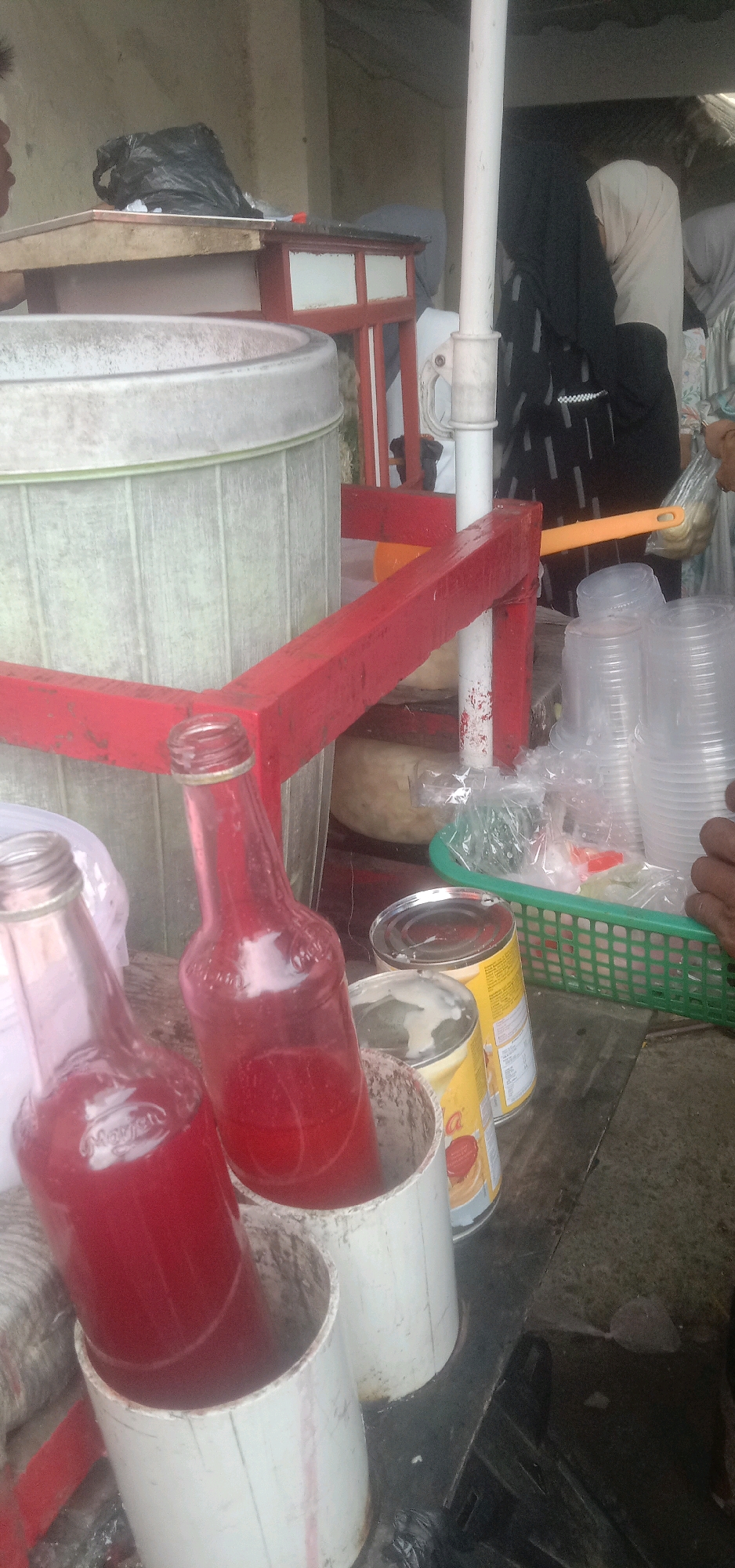This image captures a street-side beverage stand in Sukanagalih, Indonesia. Two glass bottles filled with a red liquid, likely a beverage, are prominently displayed in the foreground, each standing in a white cylindrical holder. Beside them, two yellow cans with red labels are visible, suggesting ingredients or pre-packaged items. A green plastic basket holds a stack of clear plastic cups and some wrapped items, presumably for serving. In the background, a large, light-colored cylindrical container, possibly for mixing or storing drinks, is situated behind a red-painted structure. Several individuals, partially obscured, are present in the background, some wearing head coverings, indicating a public setting. The overall environment suggests an outdoor, informal commercial setup.