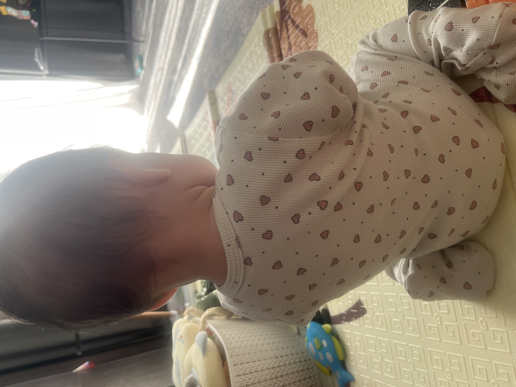 The image shows a baby from behind, sitting on a yellow playmat. The baby is wearing a white onesie with a pattern of small pink hearts and black dots. To the left of the baby, there is a white woven basket filled with light-colored stuffed toys and a blue toy fish with yellow fins and spots. In the background, sunlight streams through a sheer curtain, casting striped shadows on the floor and playmat. A dark furniture item is visible on the right. The scene appears to be indoors, likely in a living room or playroom. There is no information available to indicate the location in the city of Voronezh, Russia.