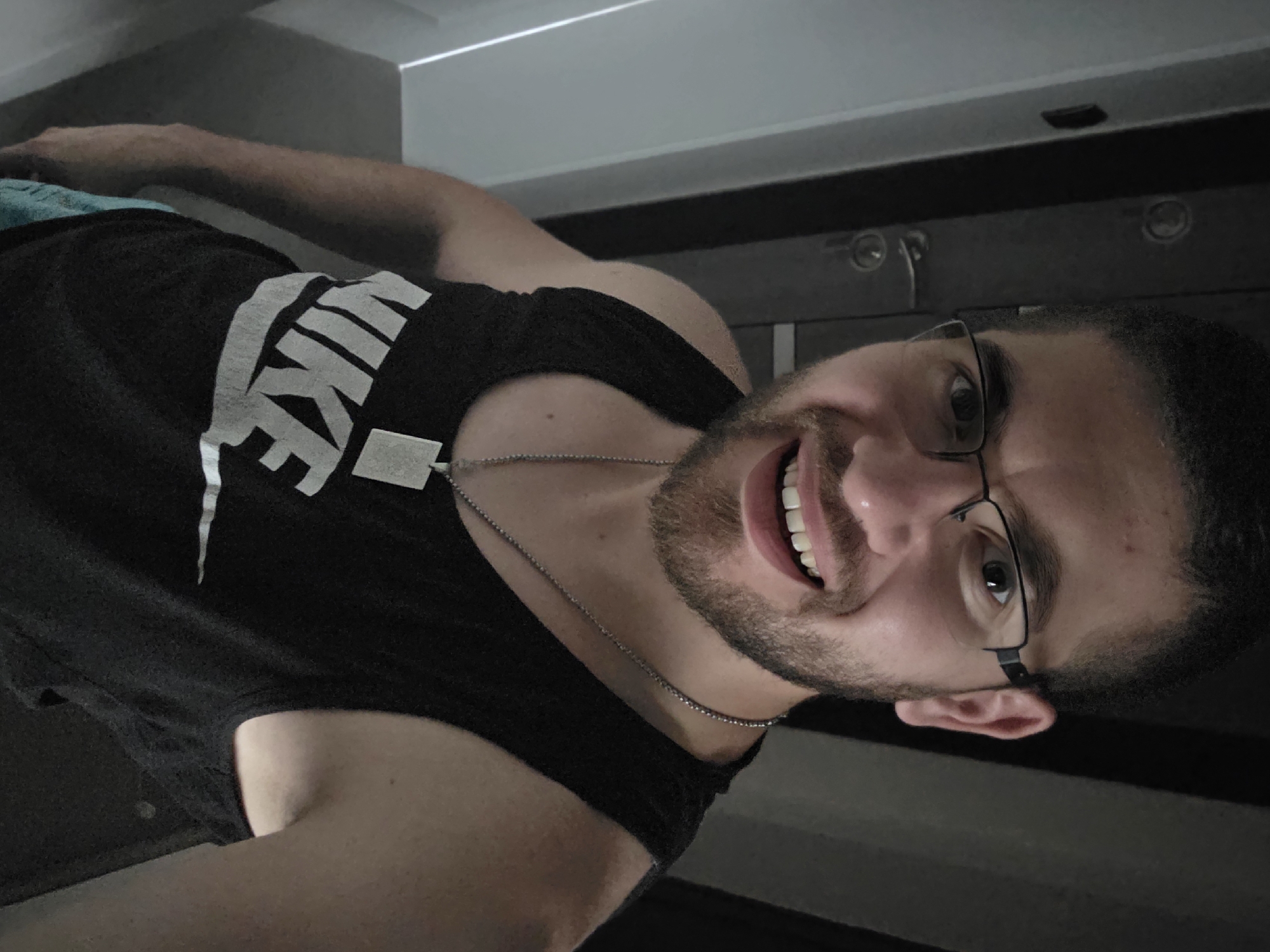 A man with dark hair and a beard is smiling and wearing glasses and a black Nike tank top with a silver chain. He appears to be indoors. The location is unknown.