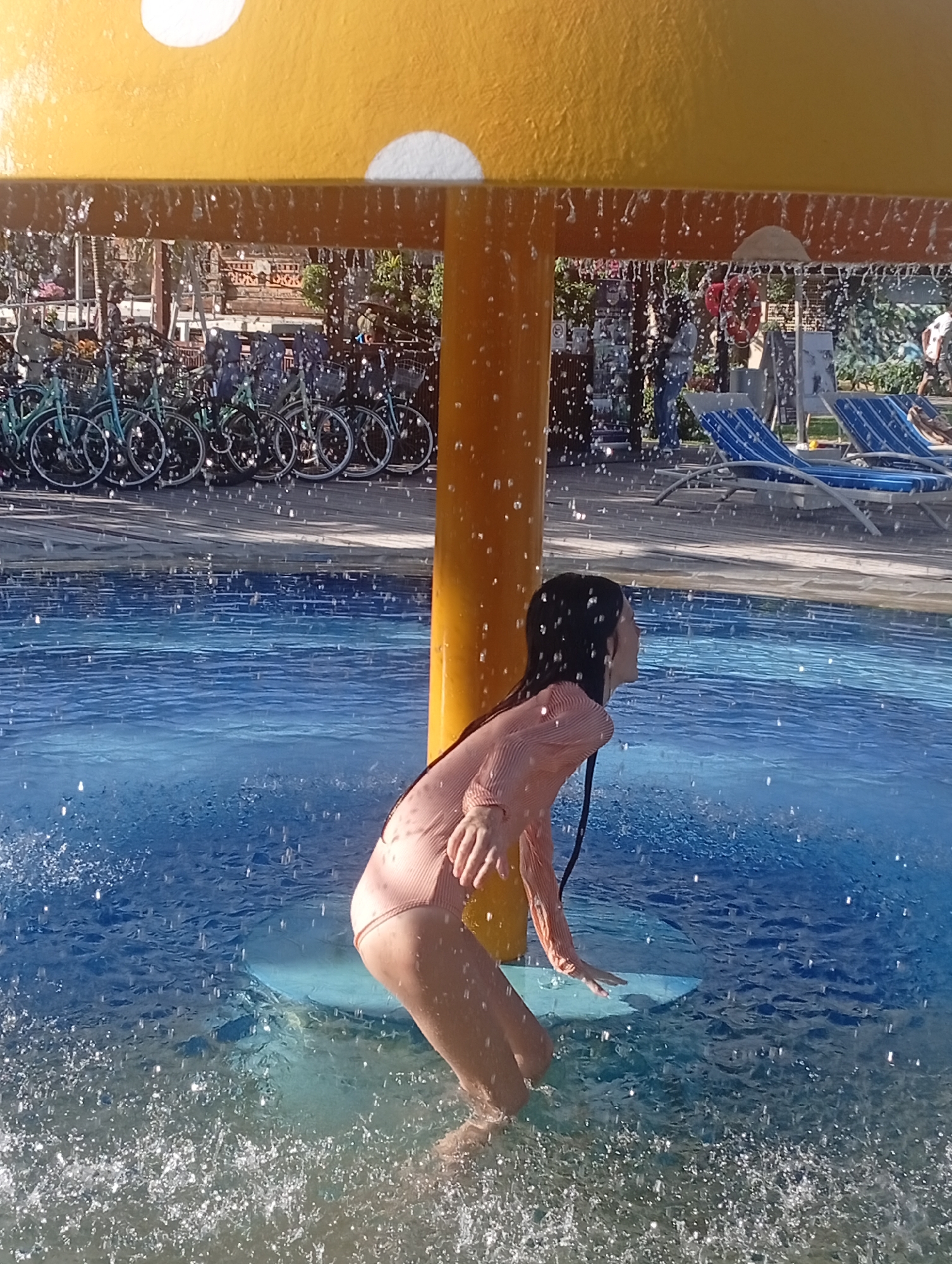 A girl in a long-sleeved pink swimsuit is standing in a shallow pool under a yellow mushroom-shaped water feature at a water park in Denpasar, Indonesia. Water is cascading down from the mushroom’s edges, creating a curtain of droplets around her. She is looking towards the right of the frame with her hand outstretched, seemingly interacting with the water. In the background, there are bicycles parked on a wooden deck, sun loungers, and other indistinct figures. The pool has a blue gradient, and the light suggests a sunny day.