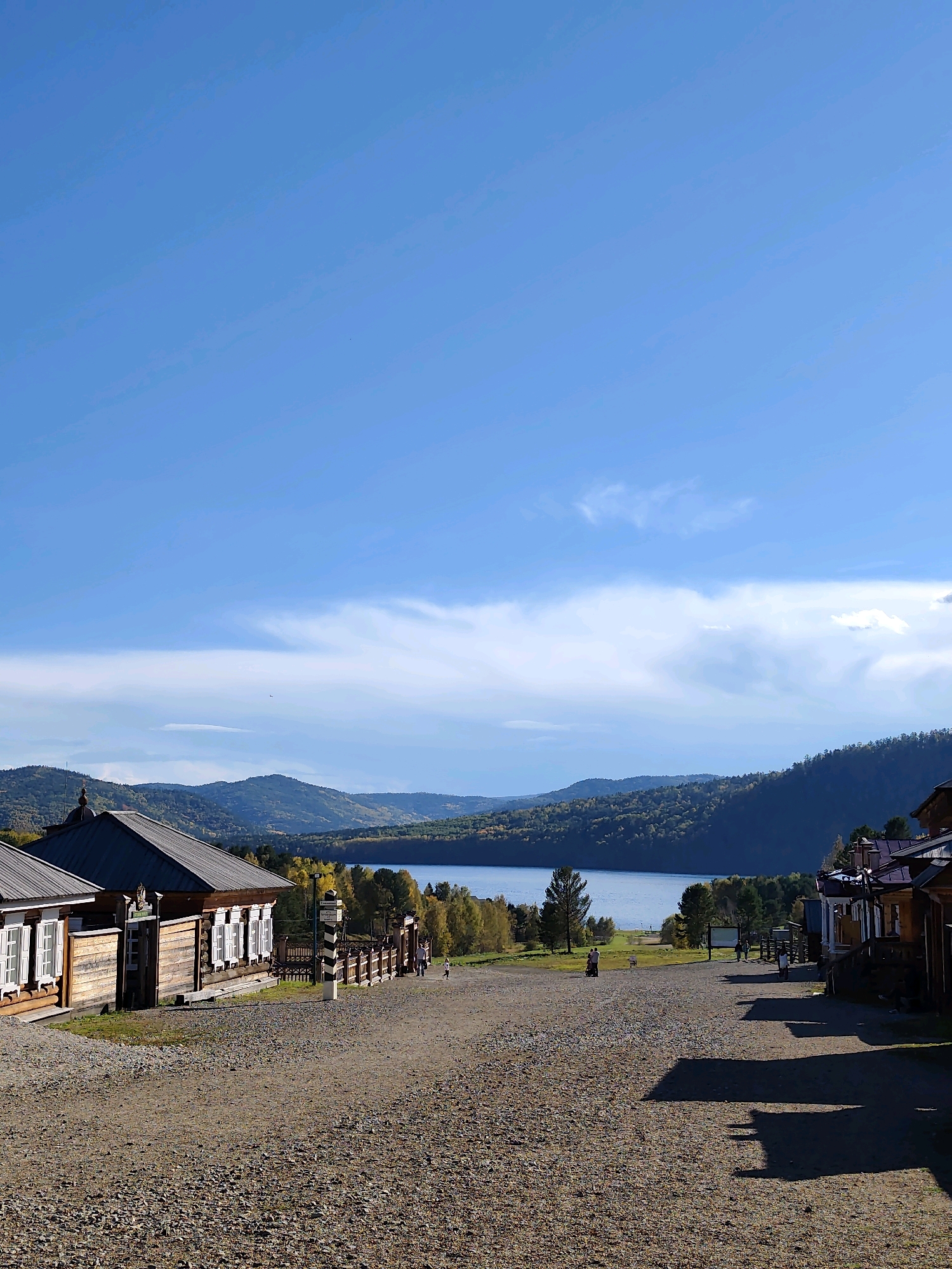 The image depicts a street scene in the city of Taltsy, Irkutsk Oblast, Russia, on a clear, sunny day. The foreground is dominated by a gravel road with scattered pebbles and dark shadows cast by buildings on the right. On the left side of the road, traditional wooden Russian houses with white-trimmed windows are visible. A fence with vertical posts and horizontal railings runs along the right side of the road, separating it from a grassy area with a few scattered trees.
In the midground, several people are walking along the road and in the grassy area. A tall, striped border post stands near the fence. Beyond the road and the grassy area lies a tranquil lake, reflecting the blue sky. The lake is surrounded by rolling, tree-covered hills. The trees on the hills exhibit autumnal colors, with yellow and orange foliage interspersed with green. The sky is a vibrant blue with scattered white clouds, some of which are wispy and stretched across the horizon. The overall impression is one of a peaceful, historical village nestled in a natural landscape.