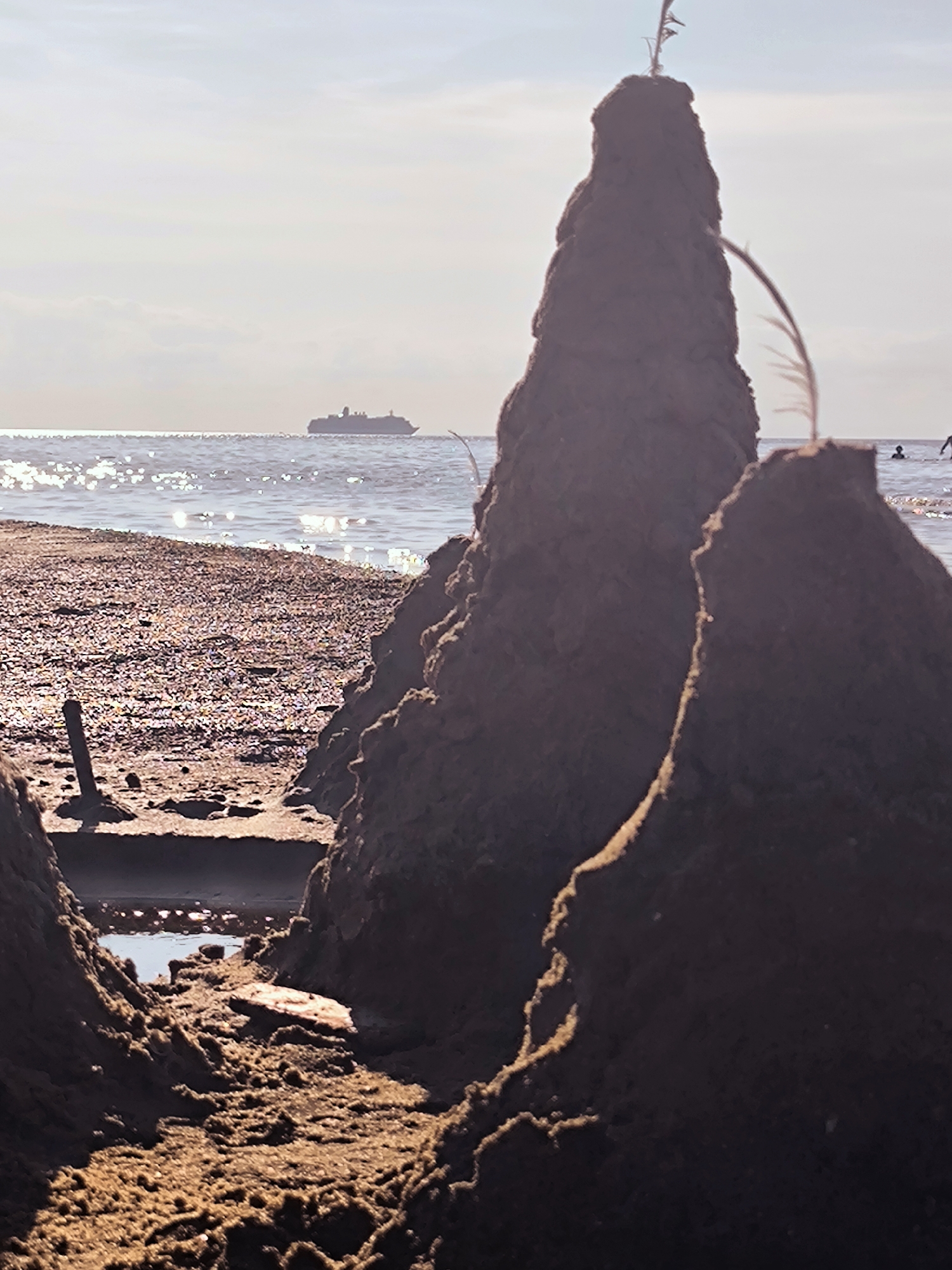 The image depicts a beach scene with a sandcastle in the foreground. The sandcastle is a tall, conical structure made of dark sand, with a smaller, partially formed structure to its right. A thin, straw-like object is stuck in the top of the main sandcastle, and another, curved, feathery object arches over the right-side structure. In the middle ground, the sandy beach transitions to the water, which is dappled with sunlight. In the distance, a large ship is visible on the horizon, sailing on a calm sea under a hazy, light-blue sky. There are two silhouetted figures in the water on the right side of the frame, one appearing to be swimming. The overall impression is a serene beach day, likely during daylight hours given the lighting conditions and the presence of the sun's reflection on the water. The scene is devoid of any specific landmarks that would indicate its location within the city of Riga, Latvia.