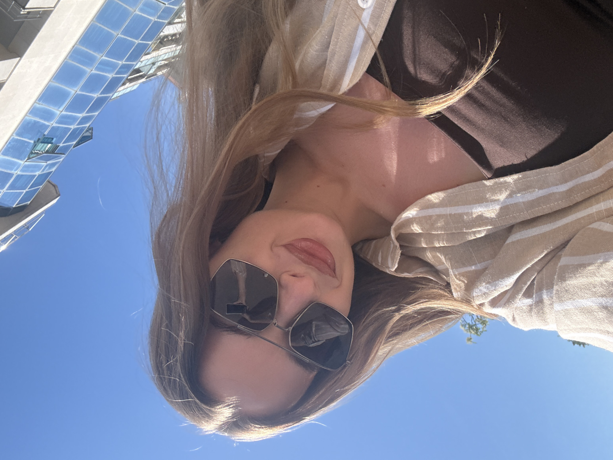 A low-angle, close-up outdoor portrait features a young woman looking upward towards the camera. The setting is bright and sunny under a clear, vibrant blue sky. Behind her, a modern building with striking blue-tinted glass windows reflects the sky and parts of the surroundings.
The woman has long, light brown hair, illuminated by the strong sunlight, with some strands falling over her shoulders and chest. She is wearing stylish, light-framed, hexagonal sunglasses, which reflect the scene in front of her, including what appears to be a phone or camera held by the photographer. Her lips bear a subtle smile and are coated with a glossy, natural-toned product.
She is dressed in a beige and white striped button-down shirt, worn open over a dark brown or black top. The sunlight creates distinct highlights and shadows on her skin, particularly on her neck and chest, emphasizing the bright conditions. The overall impression is relaxed and summery.
The scene suggests an urban or modern setting, possibly near a city center or waterfront, given the modern architecture and clear skies, consistent with the specified location of Budva, Montenegro. There is no visible text in the image.