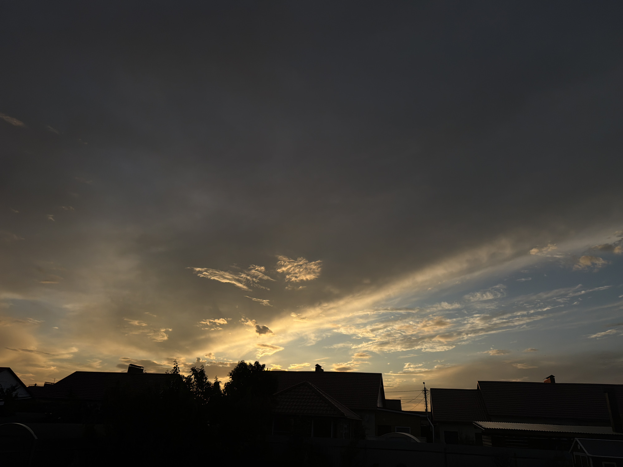 The image shows a silhouetted view of houses and trees against a dramatic sunset sky. The sky is filled with clouds, with a bright golden light illuminating them from the horizon. The tops of several red-tiled roofs are visible, along with the dark shapes of trees. The overall impression is one of a rural or suburban setting at dusk, with the natural beauty of the sunset dominating the scene. The image location is Узюково, Russia.