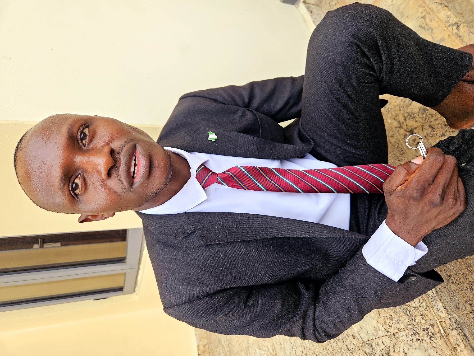 In Guzape, Nigeria, a dark-skinned man in a black suit, white shirt, and red-striped tie is captured in a portrait. He is seated on beige tiled steps, with a pale yellow wall and a window with grey panes in the background. The man is looking upwards and to his right, with his mouth slightly open. He is holding a set of keys with a silver ring in his right hand. A Nigerian flag pin is visible on the lapel of his suit jacket. The lighting suggests it is daytime.