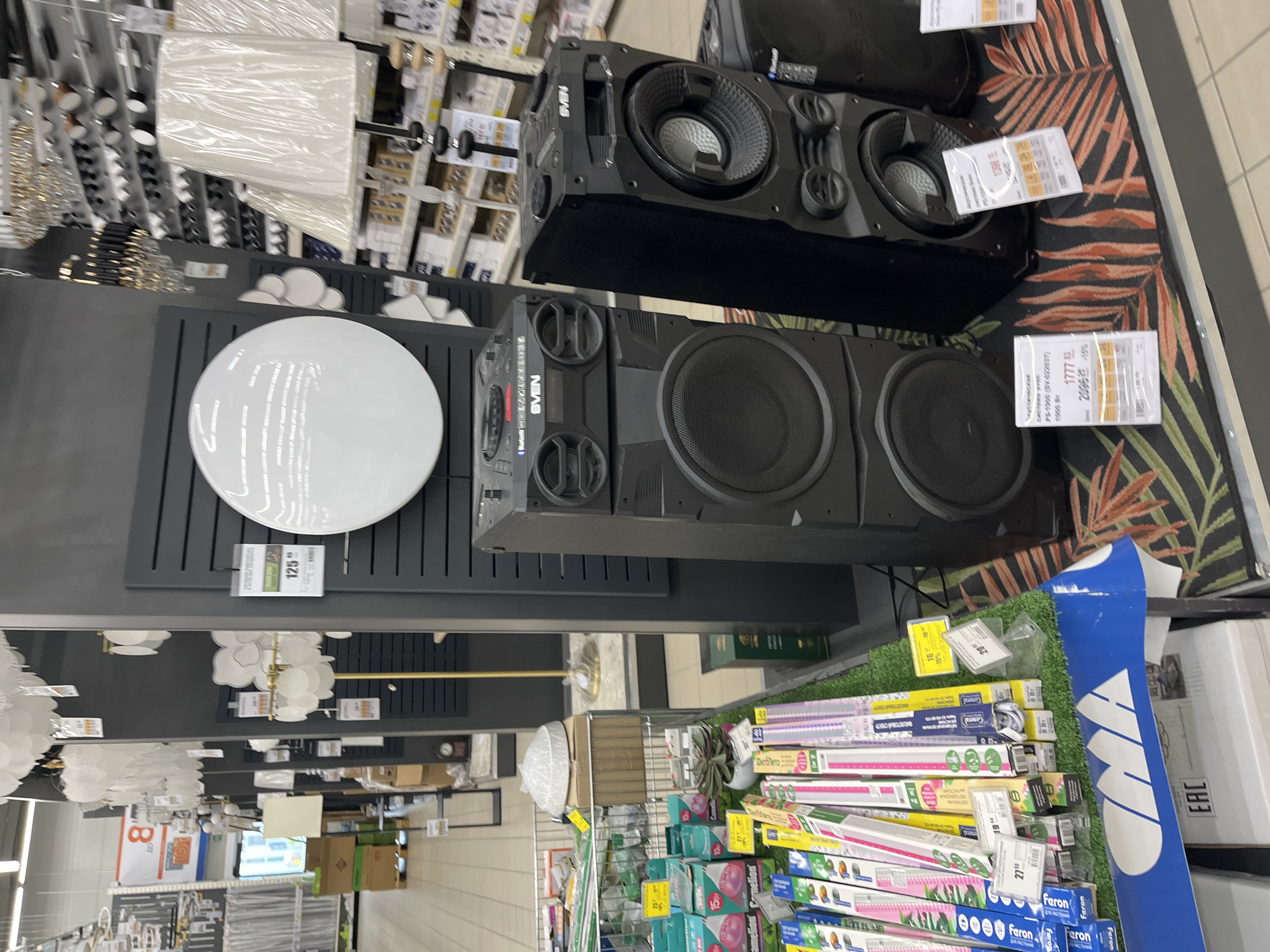 The image shows a retail display of audio equipment and lighting fixtures inside a store in Бараўлянскі сельскі Савет, Belarus. In the foreground, there are two large black speakers, branded "SVEN," stacked vertically. The right speaker is a tall, narrow unit with visible woofers, while the left speaker is a wider, shorter unit. Between the speakers and extending to the left, there is a display of fluorescent light bulbs in various packaging, some of which have price tags. A metallic cart filled with more light bulbs and other items is visible to the left. In the background, shelves are stocked with more lighting products, including chandeliers and lamps. A large, circular white light fixture is mounted on a dark wall display to the left of the speakers. A price tag for the circular light is visible, showing "125." The overall environment suggests a large retail store specializing in home goods and electronics. The flooring under the speakers has a tropical leaf pattern.