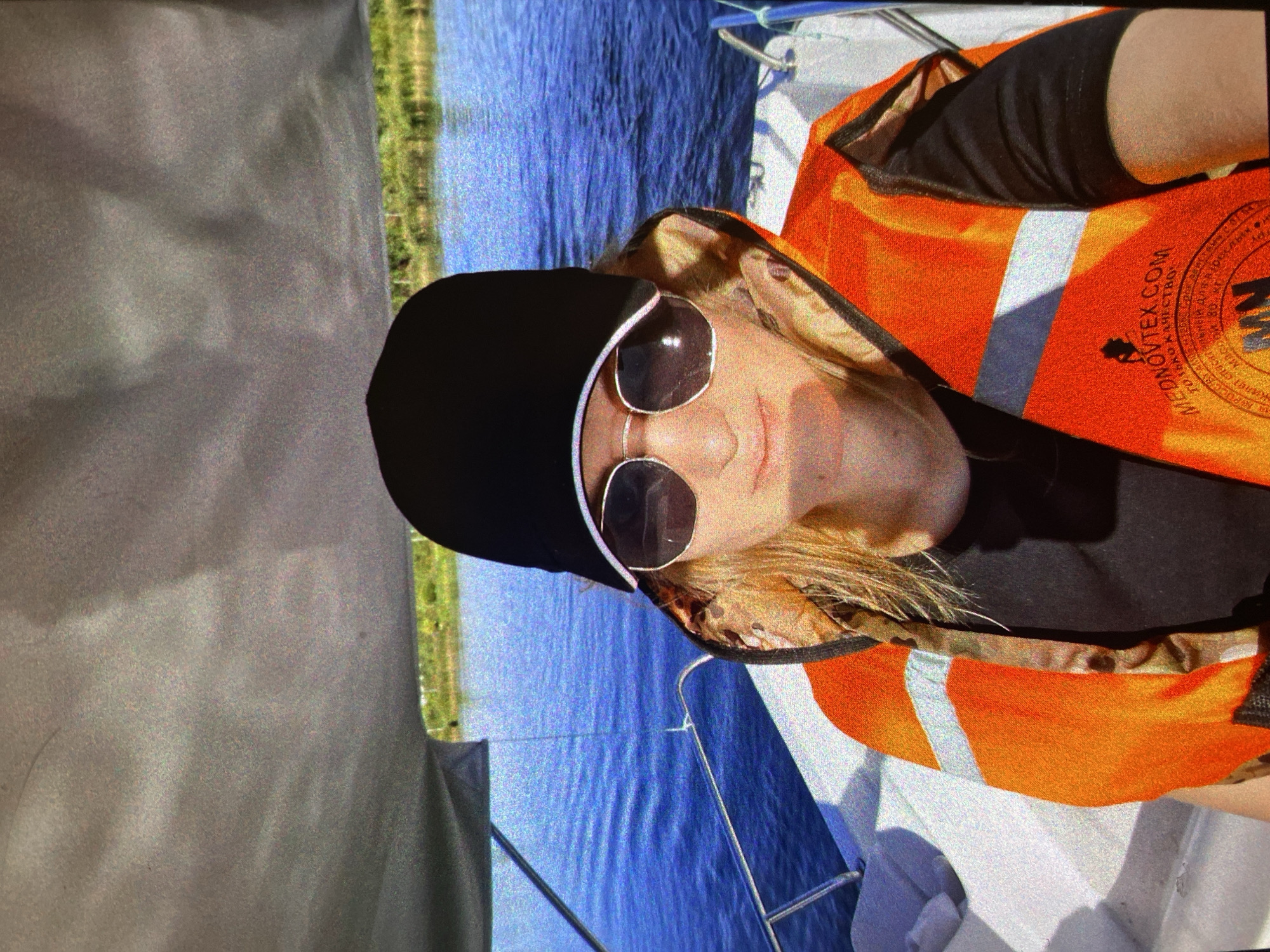 The image, rotated 90 degrees clockwise, depicts a person on a boat during what appears to be a bright, sunny day, likely in Saint Petersburg, Russia. The individual has blonde hair and is wearing dark, round-framed sunglasses, a black beanie or hat, and a black long-sleeved shirt. Over the shirt, they wear a bright orange life vest with reflective white stripes and black trim.
The person's face is partially obscured by the sunglasses and hat, but a slight smile is discernible. The life vest prominently displays text: "NOVTEK.COM" is clearly visible, along with Cyrillic words such as "МЕДПОМОЩЬ" (medical assistance) and "СПАСАТЕЛЬНЫЙ" (life-saving/rescue), indicating the vest's function and manufacturer. Other partial Cyrillic words like "ДЛЯ" (for) and "КАТЕГОРИЙ" (categories) can also be seen.
The immediate surroundings show the grey and white structures of a boat, with a metal railing visible. In the background, there is a clear blue body of water, suggesting a lake, river, or coastal area, with distant green vegetation on the shore. The sunny weather is indicated by strong highlights and shadows on the boat's surfaces. The image exhibits noticeable digital noise or graininess. The person appears to be engaged in an outdoor water activity, such as boating or sailing.