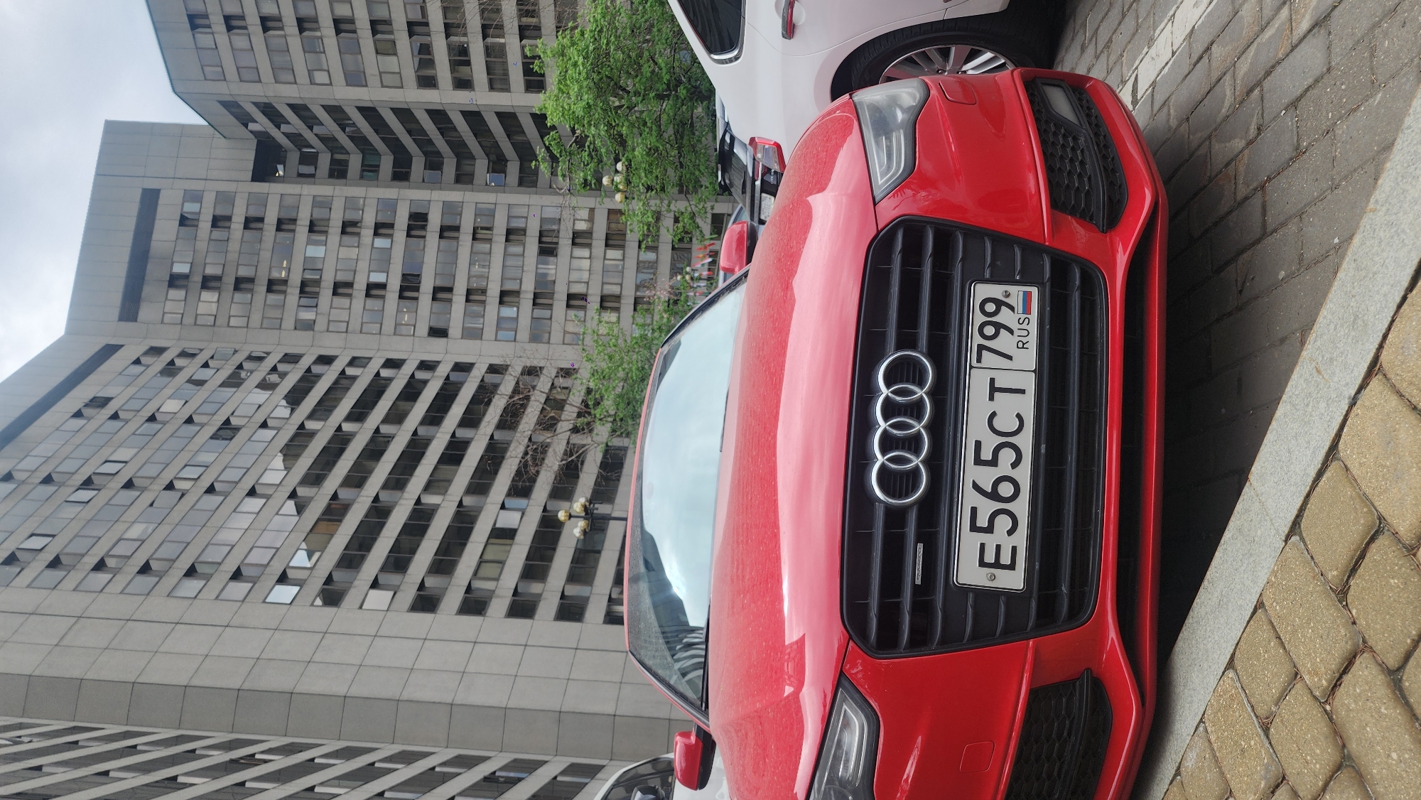 A red Audi with Russian license plate E565CT799 is parked in Moscow, Russia. The background features tall, modern office buildings with numerous windows. The scene appears to be in a business district, characterized by an urban environment. A white vehicle is partially visible behind the red Audi. The lighting suggests an overcast day.