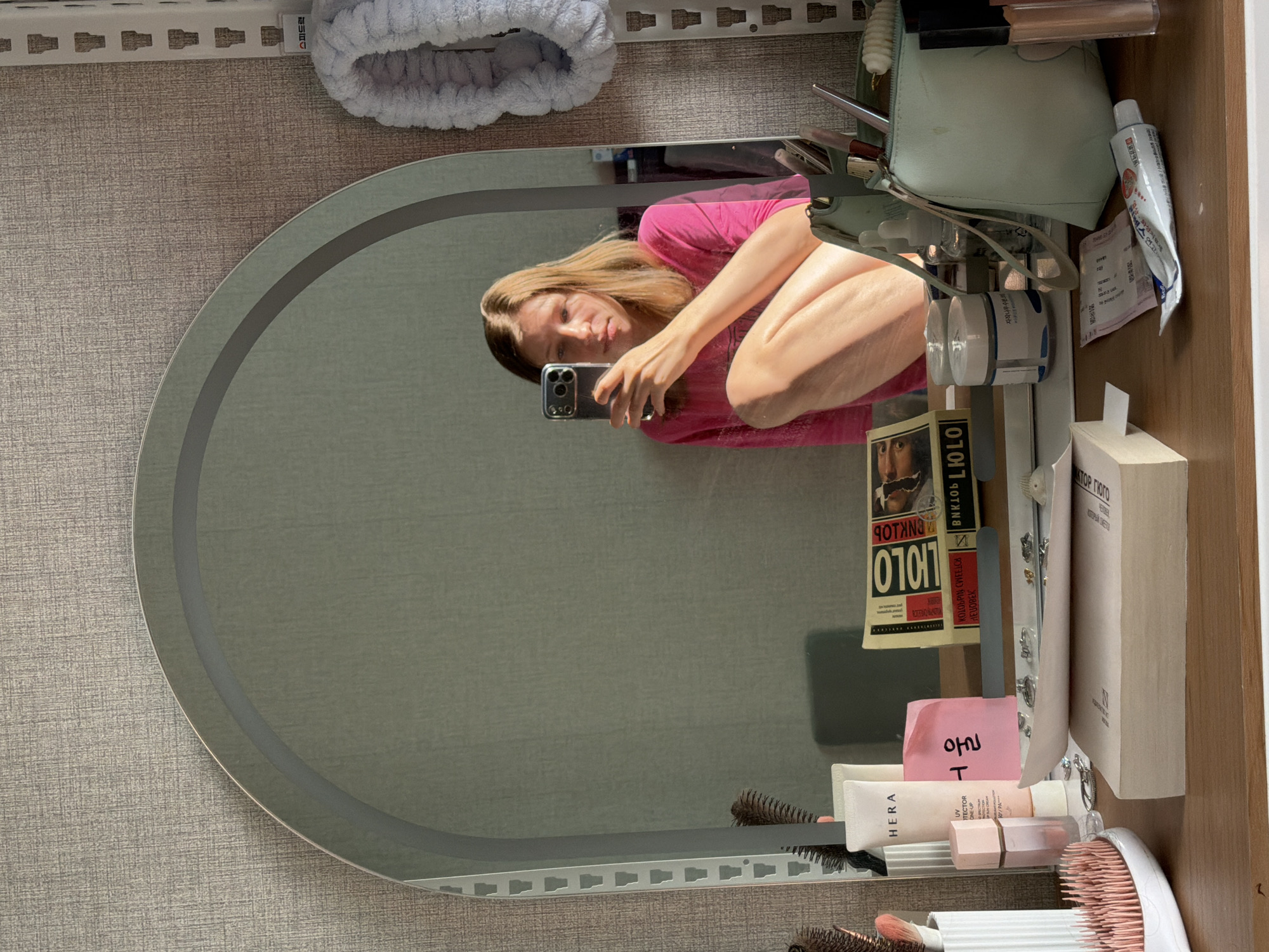 A young woman is taking a selfie in a mirror in what appears to be a bedroom or vanity area in Dongducheon-si, South Korea. She is wearing a bright pink long-sleeved shirt and is crouching, with her right arm bent and holding her phone. Her left arm is resting on her left leg. The mirror is large and oval-shaped with a light-up edge. The background behind the woman is a textured grey wall.
On the vanity in front of the mirror, there are various cosmetics and personal items. To the left, there is a white container holding makeup brushes and a pink brush. Next to it is a tube of HERA sunscreen and a small pink rectangular item. Further to the right, there are two books stacked, with one displaying the title "0101" in large letters. In front of the books is a shallow dish containing rings and other small jewelry. A larger book is placed horizontally in the foreground.
To the right of the books, there is a light blue makeup bag with various items inside, including a small white cylindrical object. A tube of white and red cream is lying on the vanity in the lower right corner. The overall scene suggests a personal grooming or dressing area.
