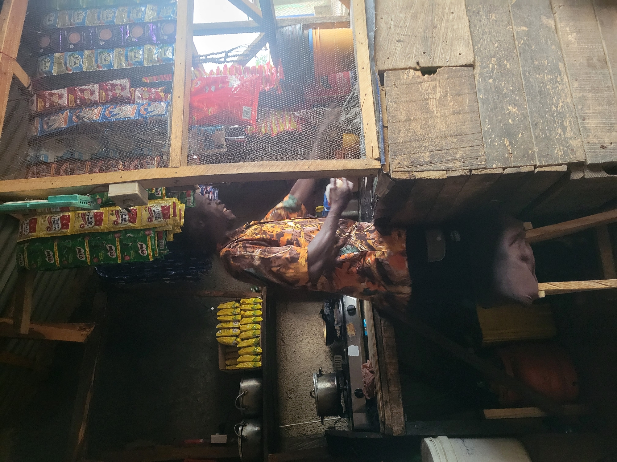 In Madalla, Nigeria, a person stands behind a wooden counter in a small shop. The shop is filled with various items, including food products displayed in plastic packaging hanging from the ceiling. There are also pots and pans on a stove, suggesting cooking is taking place or has recently occurred. The environment appears rustic, with wooden shelves and walls. The person is wearing a brightly colored shirt and is holding what appears to be a cup, possibly in the process of serving a customer or preparing a drink. The shop is dimly lit, with some natural light filtering in from an opening.