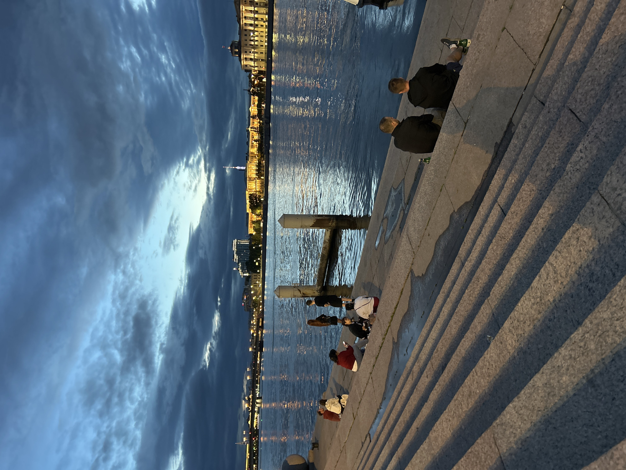 This image captures an evening scene along a waterfront in an urban setting. Several people are seated on wide, stone steps leading down to a body of water. In the foreground, two men are seated facing the water, with bottles on the steps beside them. Further up the steps, a group of individuals is gathered, some sitting and others standing. The water reflects the lights of buildings across the river, which appear to be illuminated and showcase architectural details. The sky is dark with dramatic clouds, suggesting either dusk or pre-dawn. The overall atmosphere is one of relaxed gathering and enjoying the twilight ambiance of the city.
