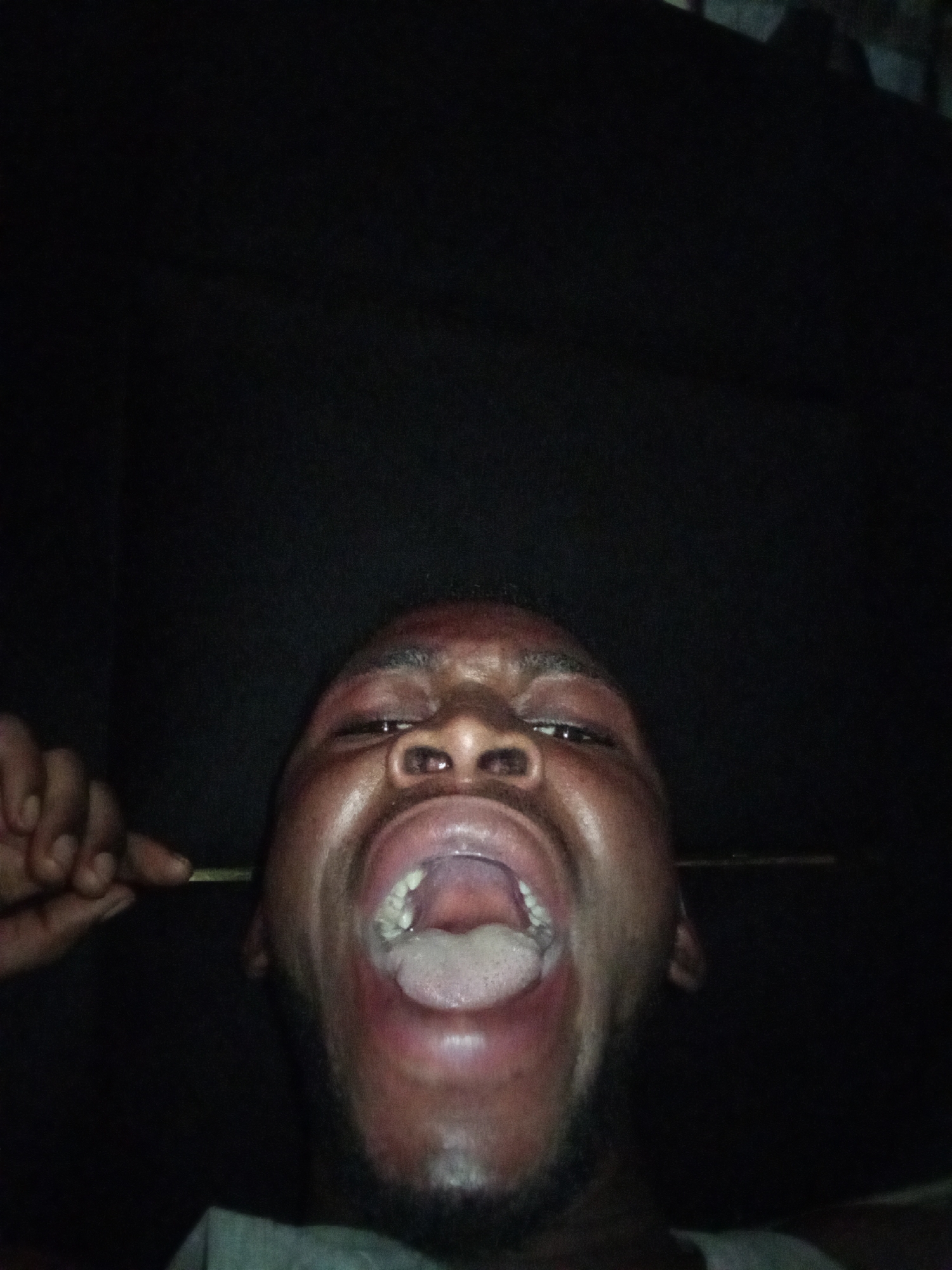 A dark-skinned man with a beard is positioned in the lower center of the frame, his face oriented towards the camera. His mouth is open wide, revealing his tongue and teeth. His right hand is visible on the left side of the frame, with his fingers extended. A thin, dark object is positioned near his ear. The background is black. The location is unknown.