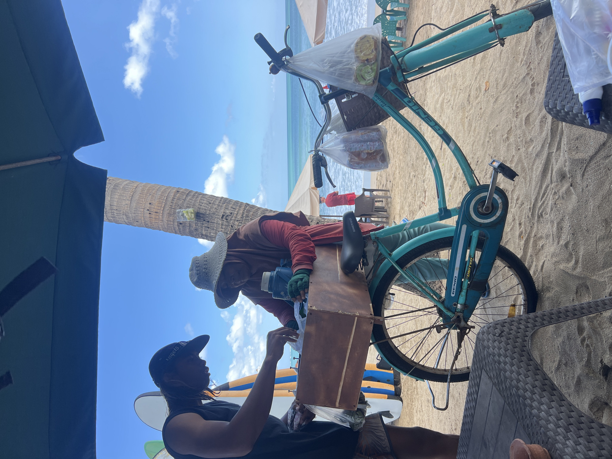 Two individuals are engaged in an exchange on a sandy beach, likely in Kuta, Indonesia, given the presence of surfboards and beach umbrellas characteristic of the area. A person wearing a dark t-shirt and a baseball cap is handing something in a plastic bag to another individual dressed in a rust-colored long-sleeved shirt, light blue pants, and a wide-brimmed woven hat. This second person appears to be operating a turquoise bicycle, which is fitted with a wooden crate over the rear wheel and handlebars adorned with two plastic bags containing what appear to be food items. The bicycle is positioned in front of a palm tree and faces the ocean, where waves are visible. The background includes more beach umbrellas and chairs. A low, woven table is in the foreground. The sky is blue with scattered clouds.