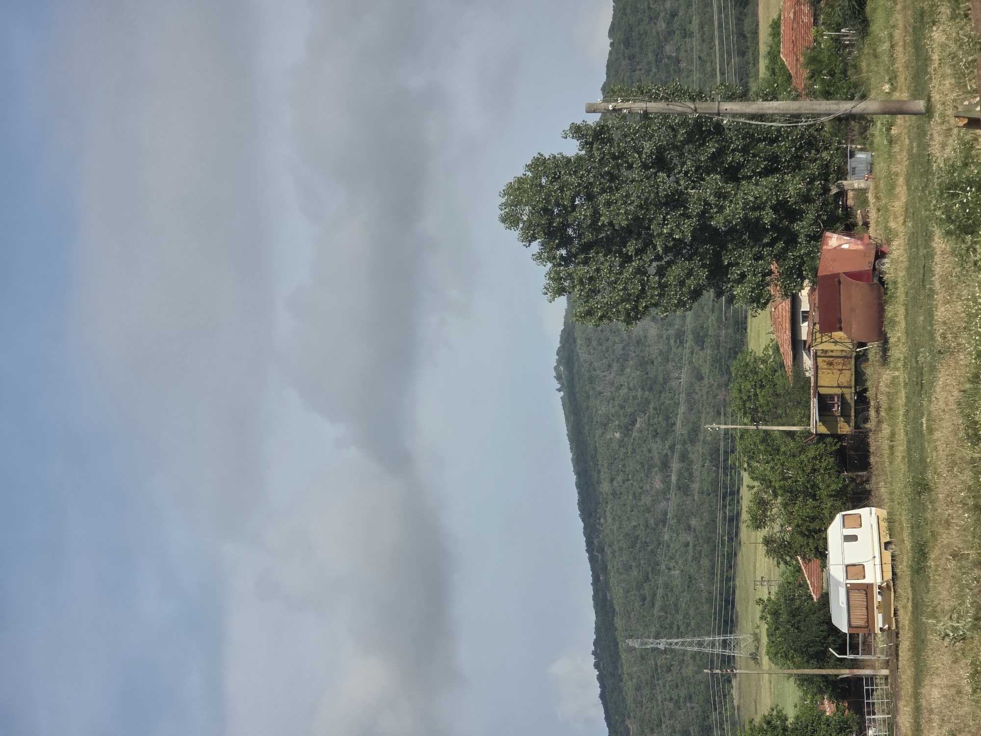 This rural landscape, set in Ivan Vazovo, Bulgaria, depicts an overcast daytime scene with a distinct natural and human-made divide. The left side of the frame is dominated by a pale, cloudy sky, indicating an overcast day with diffused light. To the right, a large, densely forested hill or mountain stretches horizontally across the mid-ground and background, covered in various shades of green foliage.
In the foreground and mid-ground, a grassy area with taller weeds and dry patches runs horizontally, separating the sky from the base of the hill. Scattered along this grassy strip are elements of human habitation and infrastructure. A prominent green deciduous tree stands out in the mid-ground, slightly to the right of the center. Several utility poles with power lines run through the scene, vertically supporting multiple parallel wires that extend towards the background.
Visible structures include several buildings with reddish-brown tiled roofs, partially obscured by trees and other objects. Among these, a white caravan or mobile home with brown-framed windows is noticeable in the lower-right quadrant. Nearby, there are two older, possibly industrial or agricultural structures: a rusty red metal object that resembles a large container or trailer, and behind it, a yellow metal trailer or truck bed. Further back and to the left of the main hill, a large metal electricity pylon stands against the forested backdrop.
The scene appears still, with no human figures or active events taking place. The overall impression is one of a quiet, rural setting. There is no visible text legible within the image.