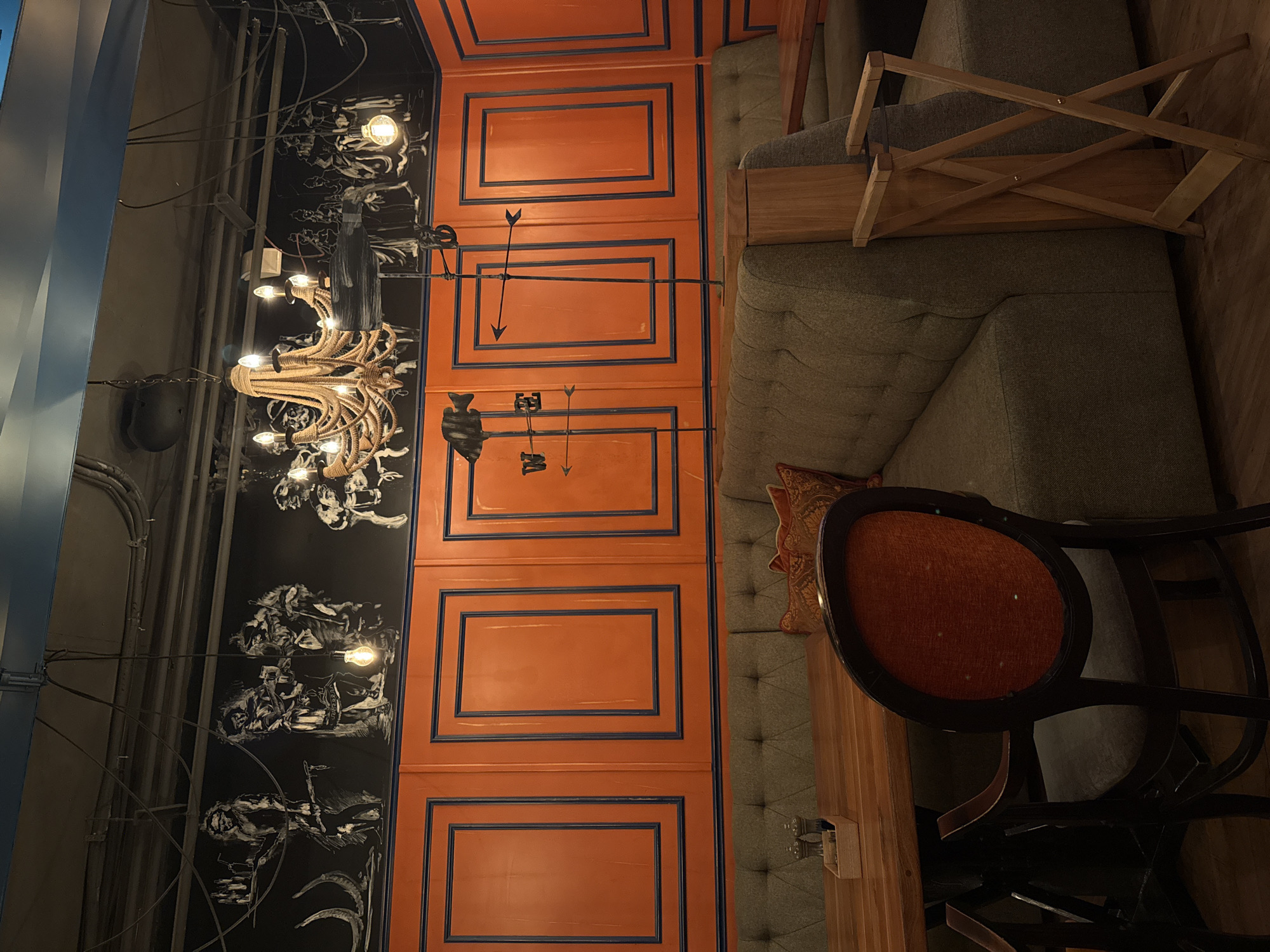 The image captures an interior space in what appears to be a restaurant or cafe in Krasnoyarsk, Russia. Dominating the left side of the frame is a wall adorned with black and white drawings, illuminated by a vintage-style chandelier. Adjacent to this wall is a section painted with orange panels framed by blue borders, featuring decorative arrow-shaped metalwork.
To the right, a plush, button-tufted sofa occupies a significant portion of the space, providing seating along the wall. A wooden table runs parallel to the sofa, and a chair with an orange fabric seat and dark wood frame is positioned next to the table. Additional wooden furniture, possibly a table or shelving unit, is visible behind the sofa.