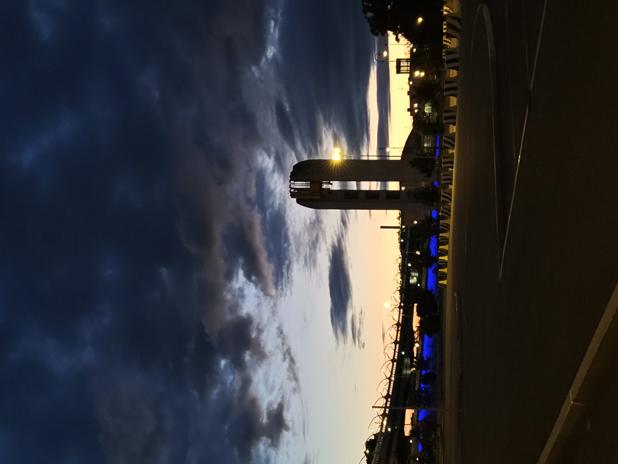 The image depicts an outdoor scene at dusk in Saint Petersburg, Russia, near the Gazprom Arena. In the foreground, there is a dark asphalt area, possibly a parking lot or open space, marked with white lines. A concrete curb runs along the left side of the foreground. Several concrete barriers with black and white diagonal stripes are arranged in a line in the midground.
Dominating the mid-right of the image is a tall, modern tower with an abstract, angular design, likely a landmark or part of a larger structure. A bright yellow streetlight is positioned near this tower, illuminating its lower section.
In the background, a long bridge structure stretches across the scene, illuminated by rows of lights that create a warm glow. Beneath the bridge, there are areas of bright blue illumination, suggesting decorative lighting. The sky above is filled with dramatic, dark storm clouds, with a band of warm orange and yellow light visible on the horizon, indicating the setting sun. Trees are visible on the far right of the image, silhouetted against the sky. The overall atmosphere is dramatic and somewhat moody due to the contrast between the dark clouds and the artificial lights. There are no people visible in the image.