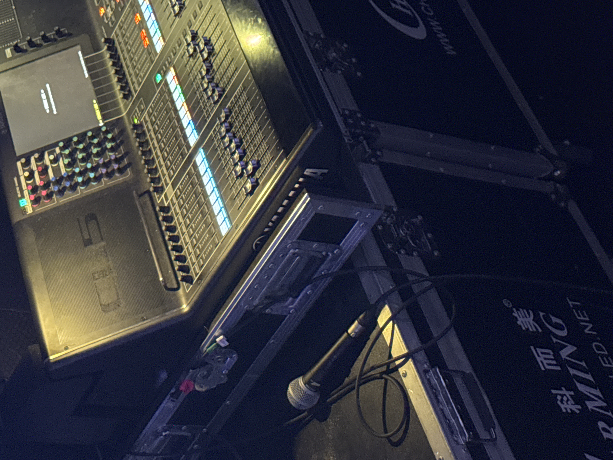 This is a close-up, high-angle shot of a black audio mixing board and a microphone on a dark surface, likely a stage or studio floor. The mixing board has numerous sliders, knobs, and illuminated buttons, with a screen displaying white text on a dark background. A silver microphone with a black handle and cord lies to the left of the mixing board. The equipment is housed within a black case with metallic accents. Part of a black case with white text and a logo is visible at the bottom. The lighting is dim, with a yellow glow emanating from the mixing board. There are no people or animals in the image, and no specific identifiable landmarks in Khabarovsk, Russia, are visible.