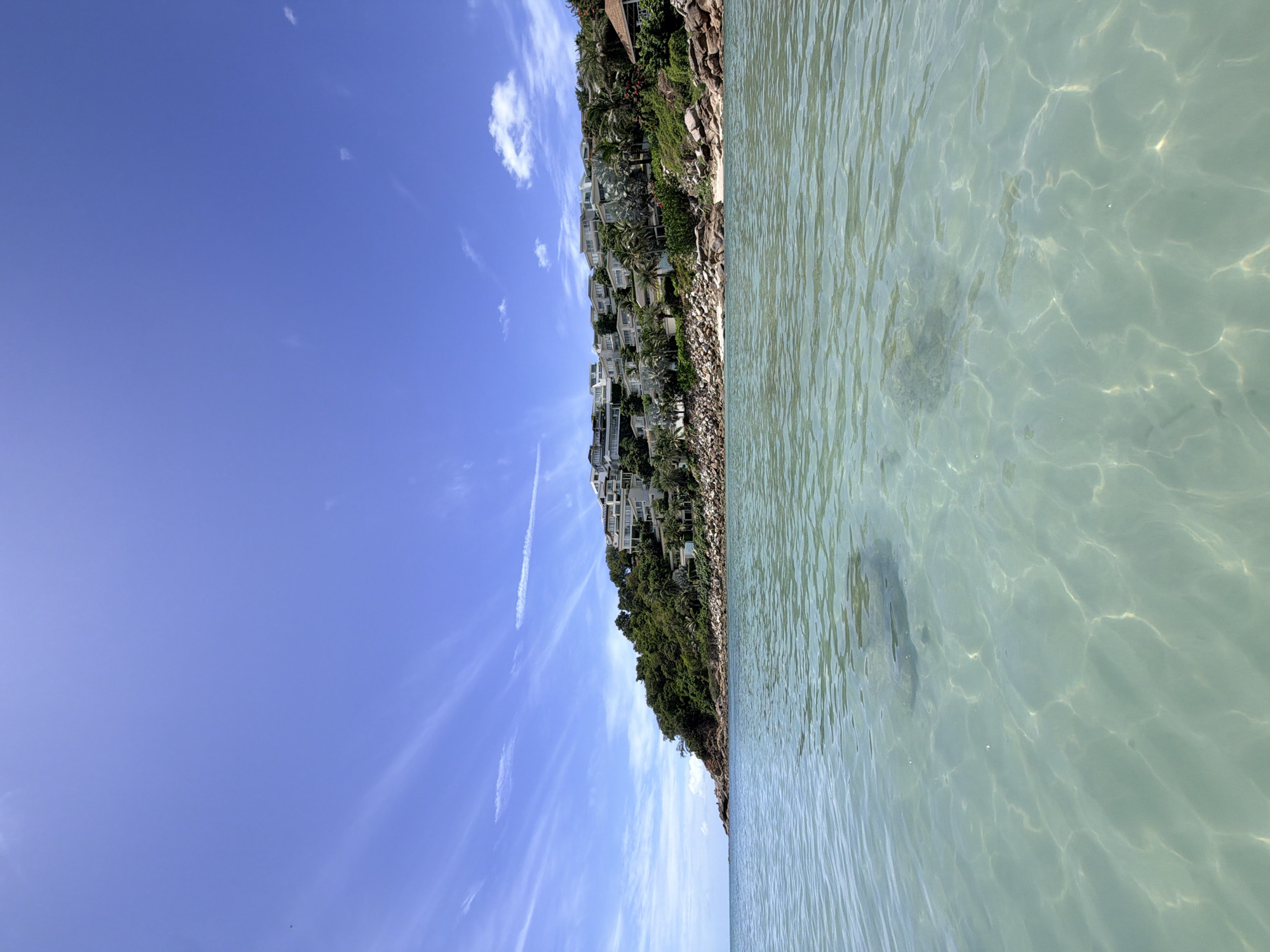 A low-angle shot captures the clear turquoise waters of the Gulf of Thailand from a perspective at or just below the surface. Sunlight refracts through the water, creating dappled patterns on the sandy bottom which is visible in some areas. The water extends towards a lush green hillside on the right side of the frame, dotted with a cluster of modern white buildings resembling villas or resort accommodations. These structures are built into the slope, with their white facades contrasting against the dense foliage of palm trees and other tropical vegetation. Rocky outcrops are visible along the shoreline, transitioning into the sandy beach in front of the buildings. The sky above is a vibrant blue, streaked with wispy white clouds that suggest a clear, sunny day. A distant headland juts out into the sea on the left, adding depth to the coastal landscape. No people are visible in the frame. The scene is characteristic of a tropical luxury resort area in Koh Samui, Thailand, likely a private development or a high-end hotel complex situated on a scenic coastal bluff.