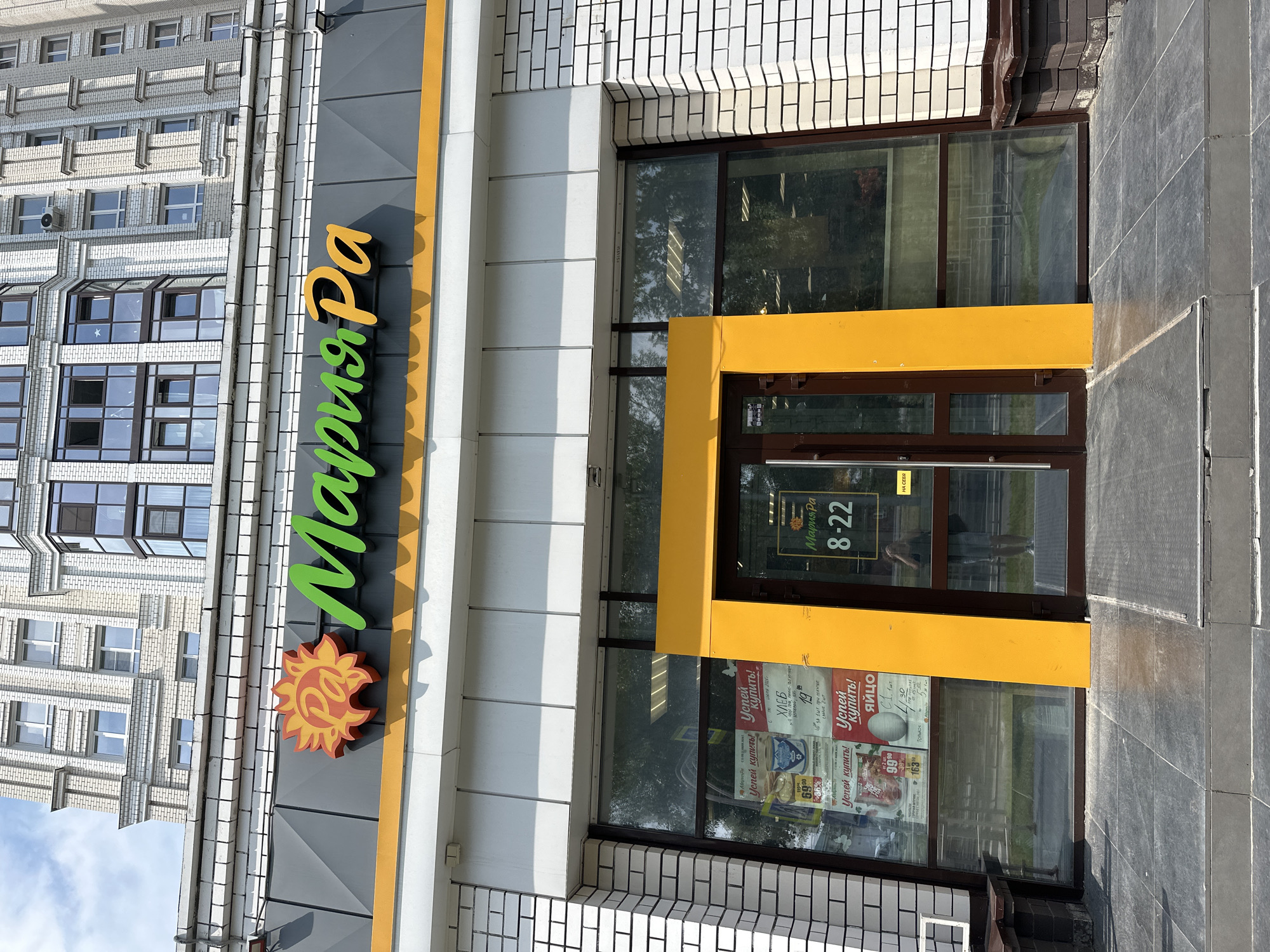 The image shows the exterior of a "Мария-Ра" (Maria-Ra) supermarket in Barnaul, Russia. The building is modern with a light-colored brick facade and large glass windows. Above the entrance, the "Мария-Ра" logo is prominently displayed with a stylized sun and green lettering. Inside the store, visible through the windows, are shelves stocked with merchandise and promotional posters. The entrance features double glass doors framed by a bright yellow structure. On the door, a sign displays the store's hours: 8-22. A person, likely a shopper, is reflected in the glass door. The area in front of the store is paved with dark gray tiles, and a small set of steps leads up to the entrance. The sky above is partly cloudy.