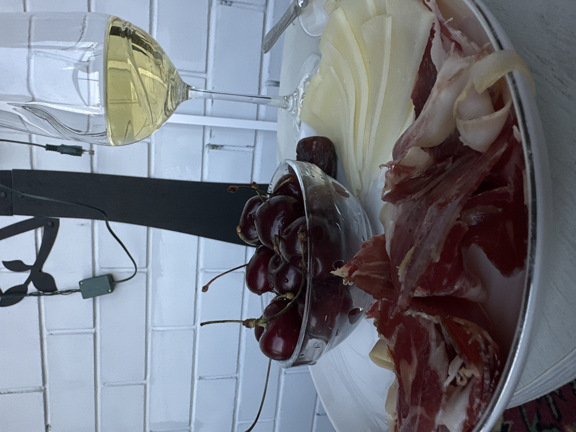 The image displays a close-up, top-down view of a spread of snacks and a drink, rotated 90 degrees counter-clockwise. Prominently featured on the left is a clear wine glass, filled approximately halfway with a light yellow liquid, likely white wine. Its delicate stem is visible extending towards the right.
Below the wine glass, a dark, decorative metal element with leaf-like patterns spans horizontally across the frame. A black wire with a small grey rectangular component (possibly an LED light controller) is draped over this element.
To the right of the wine glass, a clear, round glass bowl overflows with dark red cherries, many still attached to their green-brown stems. Some cherries appear to have water droplets, indicating freshness. Further to the right, a white circular plate holds an assortment of food. One section features neatly arranged, thinly sliced pale yellow cheese. Adjacent to the cheese, a generous serving of thinly sliced cured meat, predominantly reddish-brown with white fat marbling, is presented. This meat resembles prosciutto or a similar ham.
The background consists mainly of white rectangular tiles, suggesting a clean, possibly kitchen or patio, setting. A small portion of a patterned rug or fabric, with red and green tones, is visible at the very bottom right. The overall impression is a prepared selection of refreshments, indicating a leisurely moment or a gathering. No people are visible, and there is no discernible text within the image. The lighting appears bright and even.