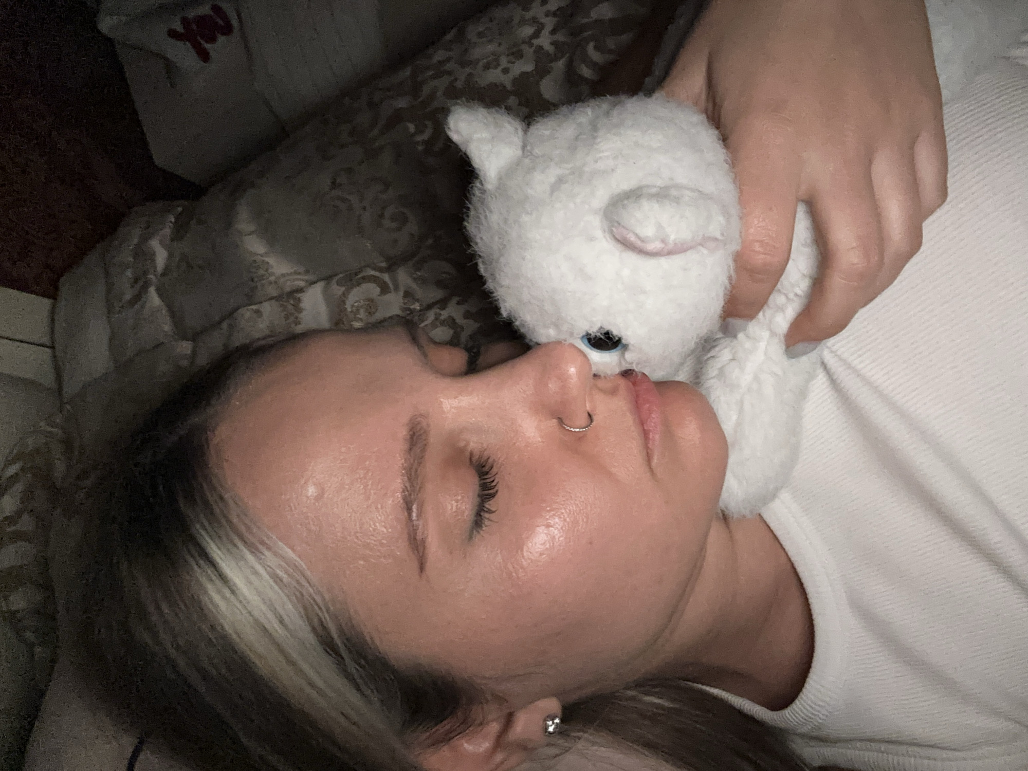 A woman is lying down, holding a white stuffed animal. She is wearing a white shirt and has a nose ring and an earring. The location is implied to be Esenyurt, Turkey. The background includes a patterned pillow and a dark area, possibly indicating a bedroom. The scene focuses on the woman's face and her interaction with the stuffed animal.
