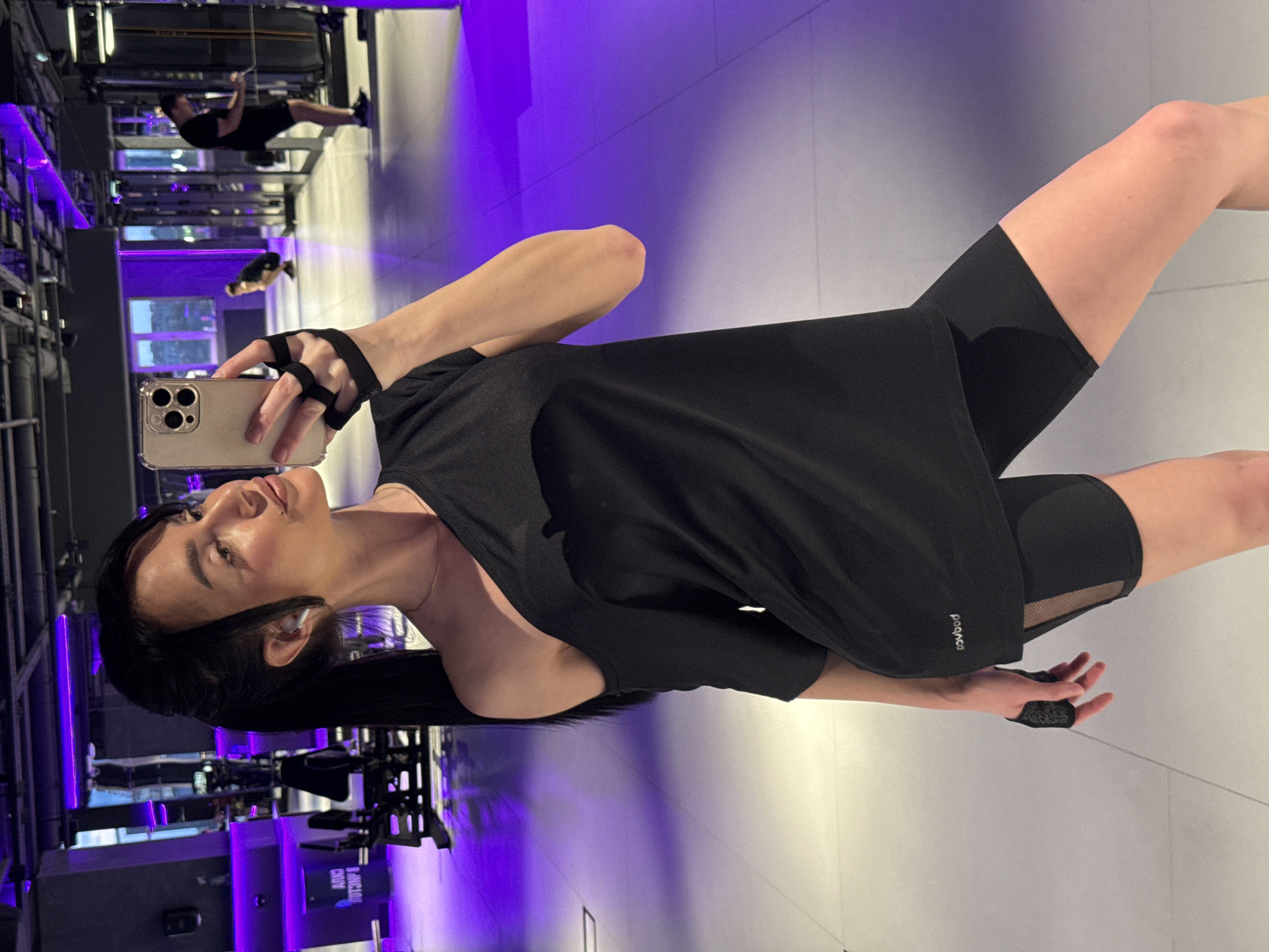 The image captures a young woman taking a selfie in a modern gym located in Krasnoyarsk, Russia. The woman, positioned prominently in the foreground, is holding a light-colored smartphone with a clear case and triple camera setup. She has dark hair styled in a ponytail with some strands framing her face, and white earbuds are visible in her ears. She wears a black athletic top and matching black cycling shorts featuring sheer mesh panels on the thighs. Black fingerless workout gloves cover her hands. Her expression is calm and she looks subtly upwards towards the camera lens.
The background reveals a sleek gym interior with a futuristic aesthetic. Striking purple neon lighting illuminates the walls and accents the structural elements of the gym, contrasting with the lighter-toned floor and other wall sections. Various pieces of gym equipment, including metal racks and exercise machines, are visible. A large reflective surface, likely a mirrored wall or glass partition, reflects the gym space, showing two male figures. One man is actively engaged in exercise, possibly performing pull-ups or dips on a piece of equipment, while another figure is seen further in the distance.
Visible text includes a small white logo on the lower right side of the woman's black top, which clearly reads "POCACO." The overall setting suggests a vibrant and contemporary fitness environment.