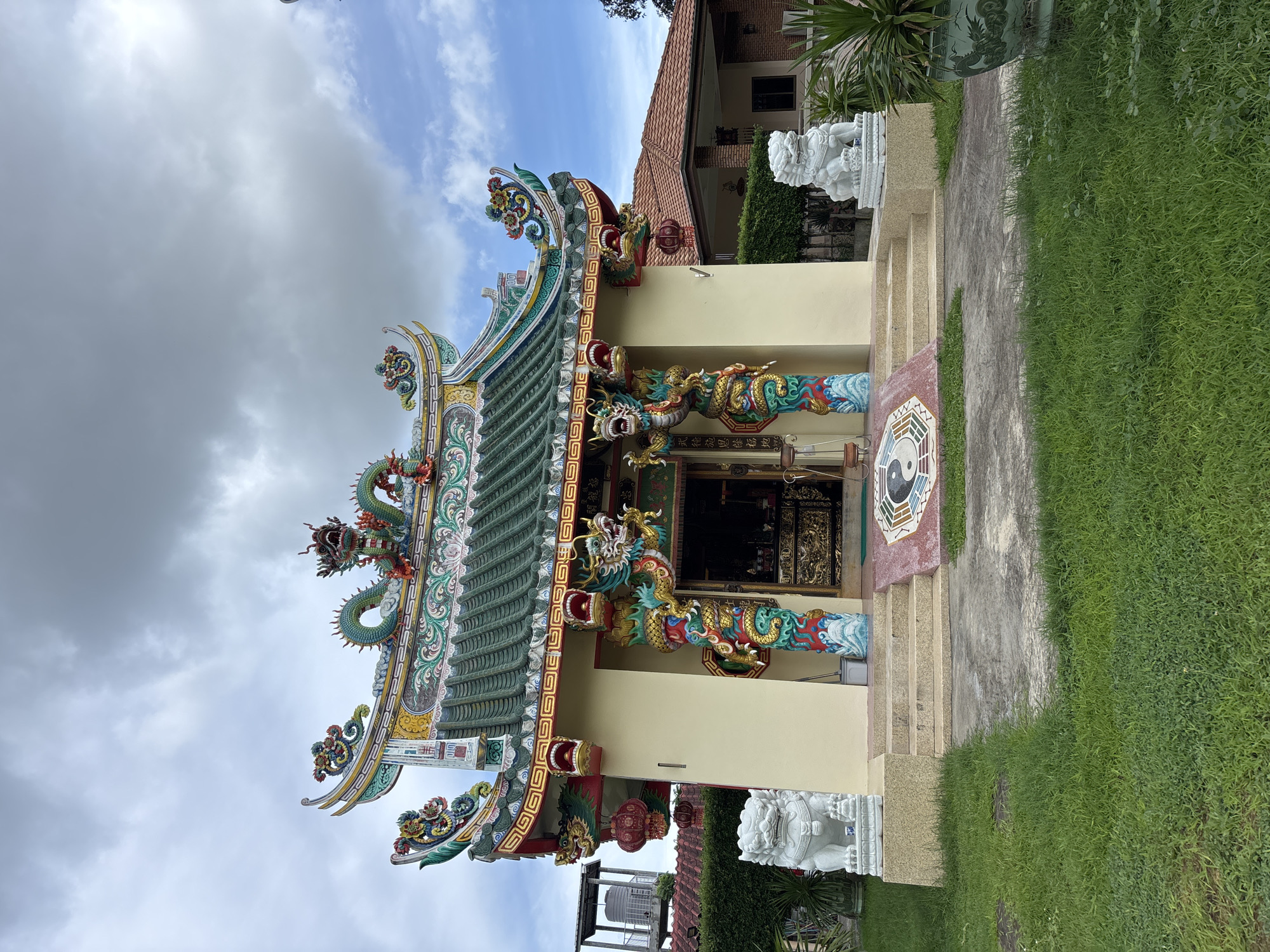 This image depicts a small, ornate Chinese temple located in Chalong, Thailand. The temple features a green, multi-tiered roof adorned with dragon sculptures and decorative elements. The building's facade is painted a pale yellow, with prominent pillars decorated with colorful dragon carvings flanking the entrance. Two white lion statues stand guard on either side of the steps leading up to the temple. In front of the steps, a circular mat displays a yin and yang symbol. The temple is situated in a grassy area with some surrounding vegetation and a residential building visible in the background. The sky is partly cloudy.