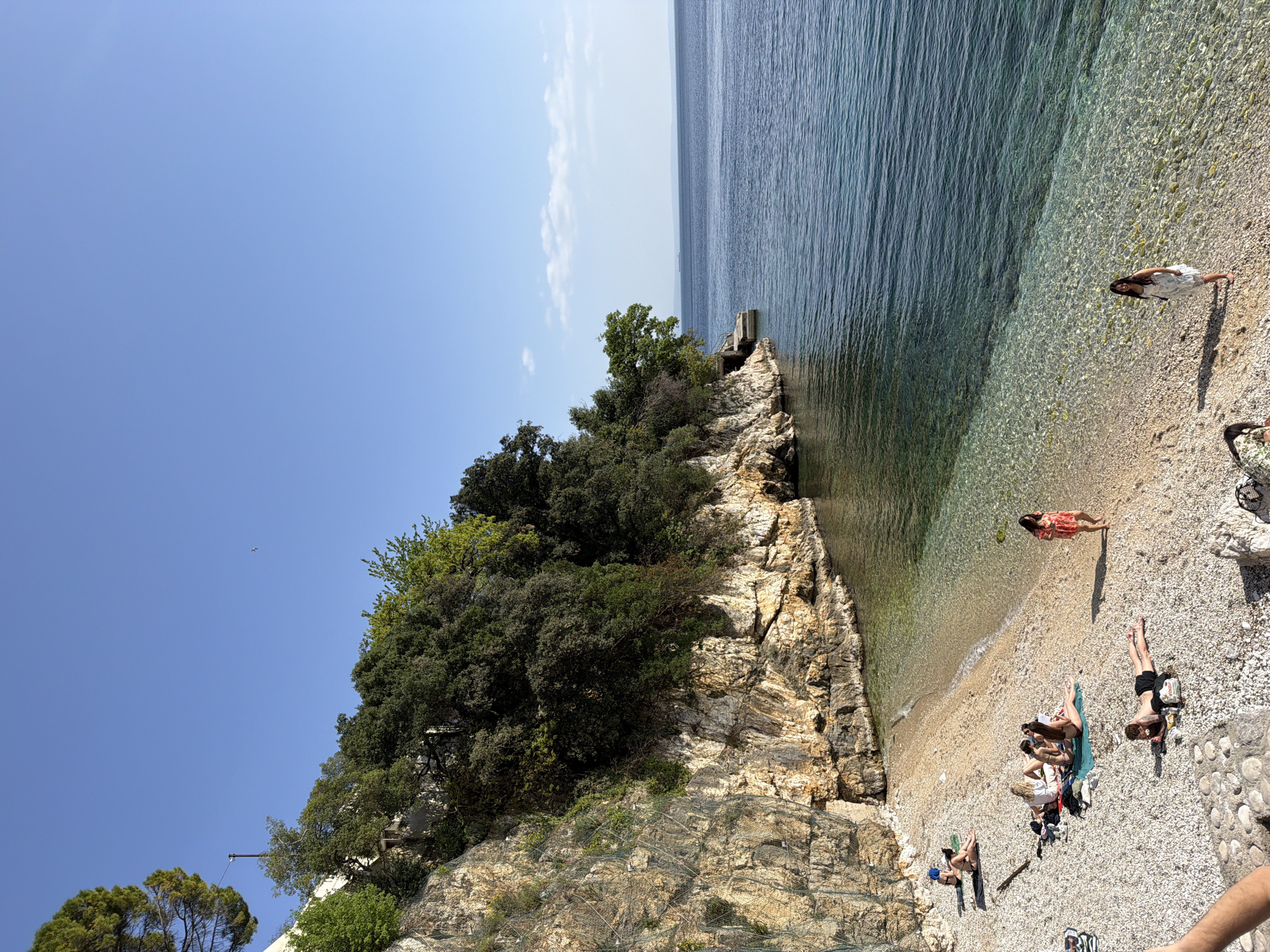 The image captures a vibrant, sun-drenched scene at a pebbly beach in Grad Rijeka, Croatia. The clear blue sky dominates the upper portion, meeting a calm, turquoise sea that stretches towards the horizon on the right. A prominent, rugged cliff face, heavily adorned with lush green trees and vegetation, anchors the left side of the frame, descending to the beach. Evidence of rockfall netting is visible on parts of the cliff, suggesting maintenance for safety.
Several individuals are present on the beach, engaged in leisure activities. A woman in a white dress stands near the water's edge on the right, looking out at the tranquil sea. Closer to the base of the cliff, another woman in a red-patterned dress lies relaxed on the pebbles, appearing to sunbathe, with a green object (possibly a fruit or small item) nearby. In the lower-left section of the beach, a small group of people are gathered, resting on towels and mats, further enjoying the sunny weather. One person lies face down, while others are seen sitting or reclining, with a dark bag visible beside them.
The water is exceptionally clear near the shore, allowing the light-colored pebbles on the seabed to be distinctly visible. A small wooden structure, possibly a pier or platform, extends from the cliff into the sea. The bright sunlight suggests it is mid-day or early afternoon, contributing to the overall atmosphere of relaxation and natural beauty. No visible text is present in the image.