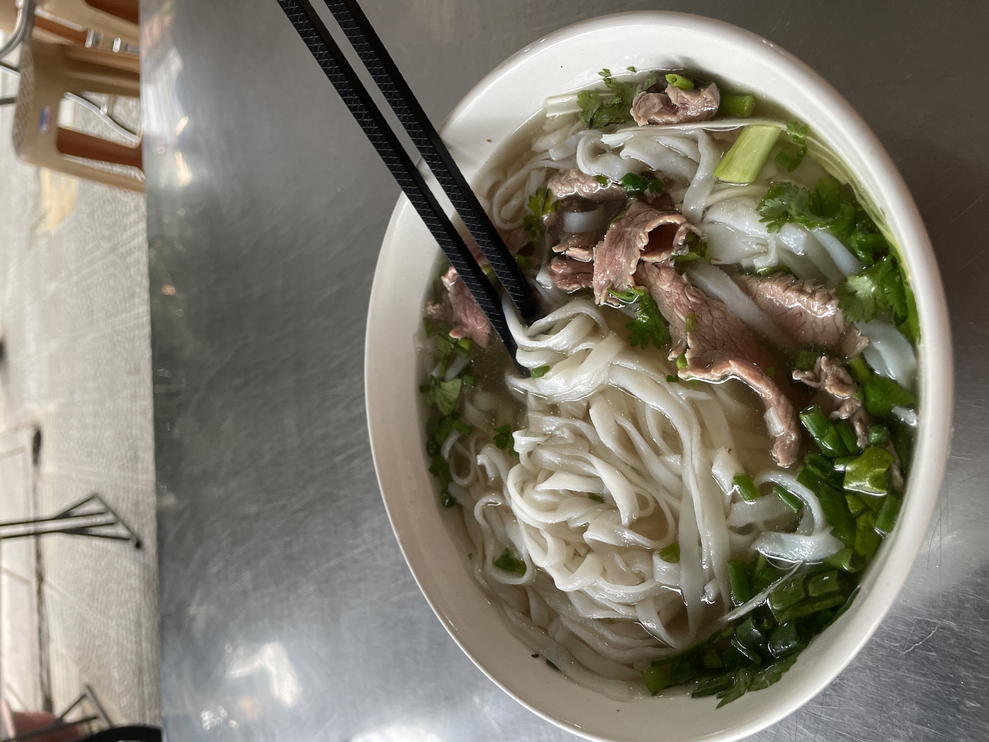 A bowl of Vietnamese pho is presented on a metal table outdoors, likely in a street-side eatery in Da Nang, Vietnam. The pho contains wide, flat rice noodles, thinly sliced beef, and is garnished with chopped green onions and cilantro. Black chopsticks rest across the top of the bowl. In the background, a textured pavement and a few plastic stools are visible, suggesting a casual dining setting. The lighting is natural, indicating daytime.