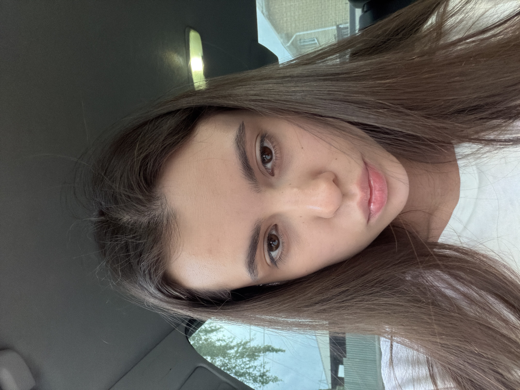 A young woman with long, straight, light brown hair is positioned in a car. She has light skin, brown eyes, and natural-looking lips. The interior of the car is visible with a dark gray dashboard and a glimpse of the side mirror. Outside the car window, a building and some greenery are partially visible. The location is not identifiable as Chaikovsky, Russia, based solely on the visual information provided. The image appears to be a selfie or a photo taken at close range.