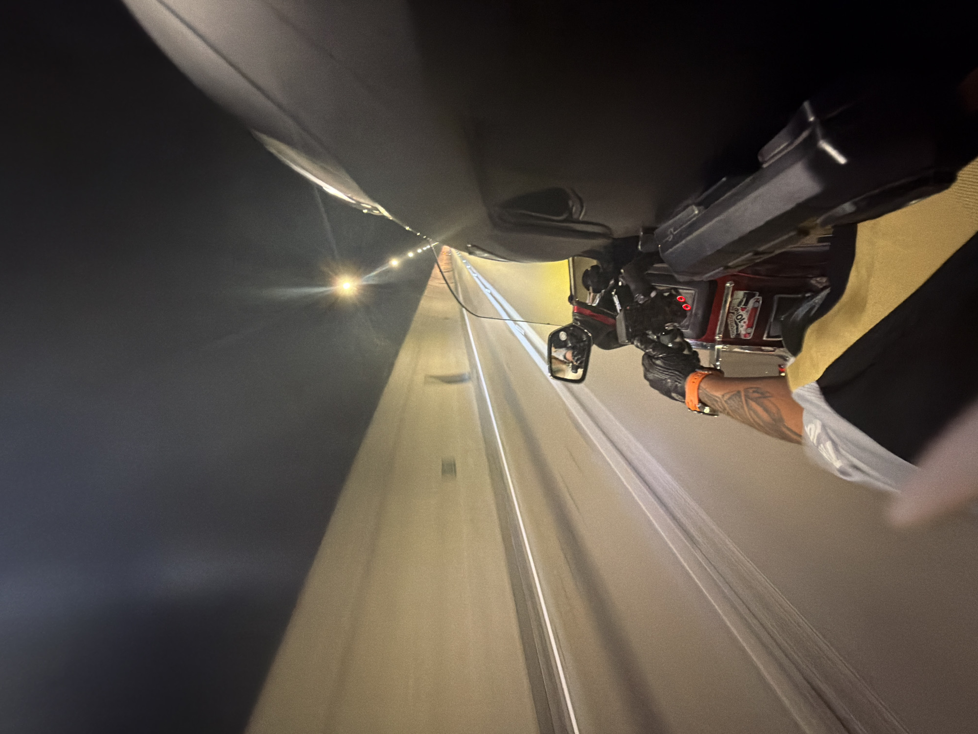 A first-person perspective shot captures a motorcycle traveling through a tunnel at night. The tunnel walls are a light beige color, illuminated by overhead lights that create streaks of light due to motion blur. A white line marks the edge of the road. In the foreground, the rider's gloved hands are visible on the handlebars, with an orange watch on the left wrist. A tattoo is visible on the rider's arm. The motorcycle's windshield and part of the rider's helmet are prominent. A rear-view mirror reflects the tunnel lights. The overall impression is one of speed and movement through an urban or suburban infrastructure. The location is identified as a tunnel in Đurmani, Montenegro.