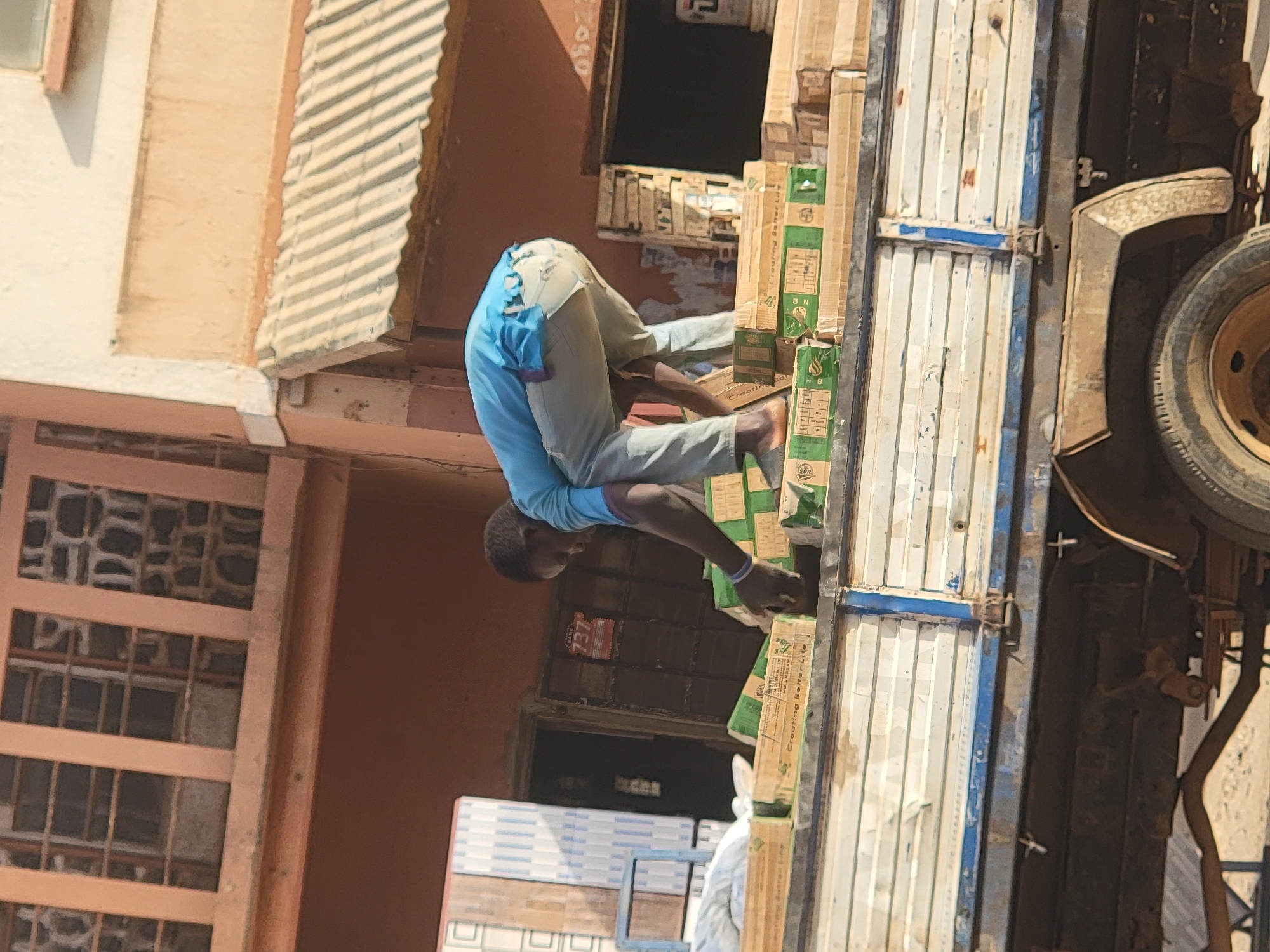 In Maiburiji, Nigeria, a man wearing a light blue shirt and light-colored pants is unloading boxes from the back of a truck. The truck has white doors with blue trim. The building to the left is brown with multiple window panes. Tiles can be seen near the doorway of the building.