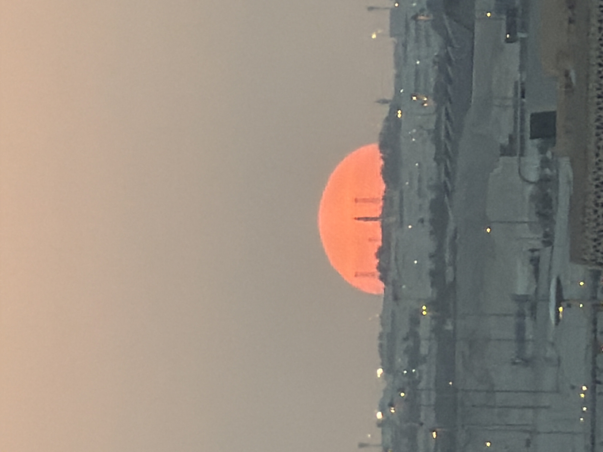 A large, glowing orange sun is setting behind a city skyline. The sun appears as a perfect, semi-circular disk, casting a warm hue across the sky. Silhouetted against the sun are several tall, industrial-looking structures, possibly smokestacks or communication towers. The city itself is spread out below, with a mix of buildings and infrastructure visible. Lights from the city twinkle in the distance, some appearing as bright dots and others as blurred streaks. The foreground shows a closer view of urban elements, including what looks like a road or paved area with streetlights. The overall impression is one of a vast, industrial landscape bathed in the dramatic light of a sunset.