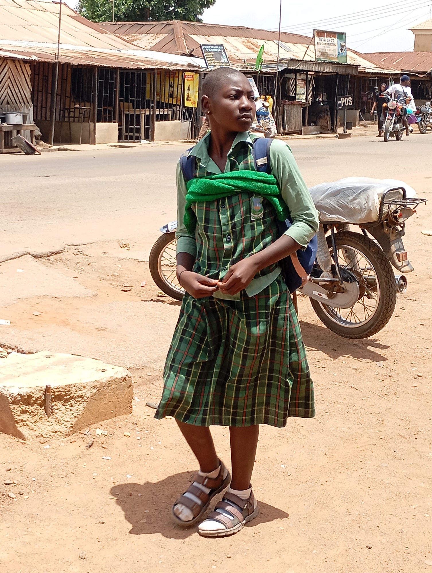 A full-shot photograph depicts a young person, likely a student, standing on a dusty roadside in what appears to be a semi-urban or rural area of Vom, Nigeria. The individual has a closely shaved head and is wearing a green plaid school uniform, which includes a long-sleeved light green shirt and a matching knee-length skirt. A blue backpack is visible on their back, and a bright green towel or cloth is draped over their shoulders. They wear open-toed sandals over white socks. A number "6" is visible on the plaid shirt near the left shoulder. The subject stands with a neutral, observant expression, looking towards the right side of the frame.
Behind the person, a motorcycle is parked, its rear section covered with a white tarp or plastic sheeting. The immediate foreground consists of sandy, uneven ground with some small rocks and a concrete block to the left. Beyond the unpaved shoulder, an asphalt road stretches across the midground.
The background reveals a bustling street scene with various single-story buildings featuring corrugated metal roofs and simple wall constructions, characteristic of shops or residences. Some buildings have barred windows. Several individuals are visible in the distance, including people on motorcycles and pedestrians. Bright daylight illuminates the scene, casting strong shadows, indicating it is likely midday or early afternoon with clear weather.
Visible text on signs in the background includes "NEW WORLD BOTTLING..." on the left, "DUTY SPOT..." near the center, and a prominent black sign displaying "POS" (likely indicating a Point of Sale service) to the right of the street. The scene captures an ordinary moment of daily life in a lively local street environment.