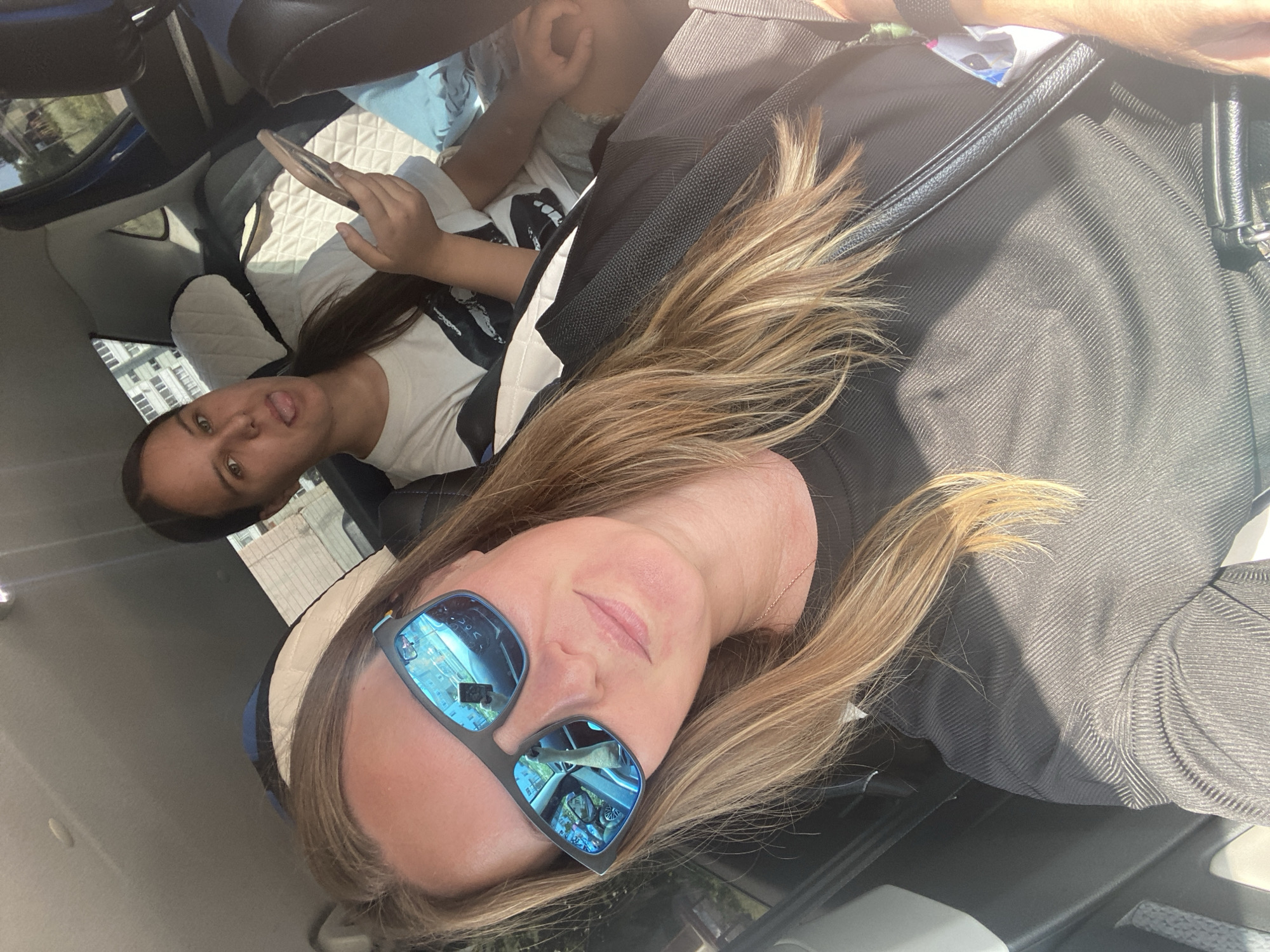 Two females are in the back seat of a vehicle, appearing to be in transit. The person in the foreground, on the left, is wearing sunglasses with blue lenses, a black ribbed shirt, and a black purse strap is visible. Her hair is blonde. She is looking at the camera, and the reflection in her sunglasses shows the interior of the car, including another person and the driver's side mirror. The second female, in the background on the right, is wearing a white t-shirt with a graphic print and is holding a smartphone. She is sticking her tongue out at the camera. The interior of the car is visible, with quilted headrest covers. Exterior elements suggest a daytime setting, with sunlight and a building visible through the car windows, possibly in a suburban or urban environment. Based on the text "Noviy, Russia" which is not in the image but provided as context, the location is assumed to be Noviy, Russia.