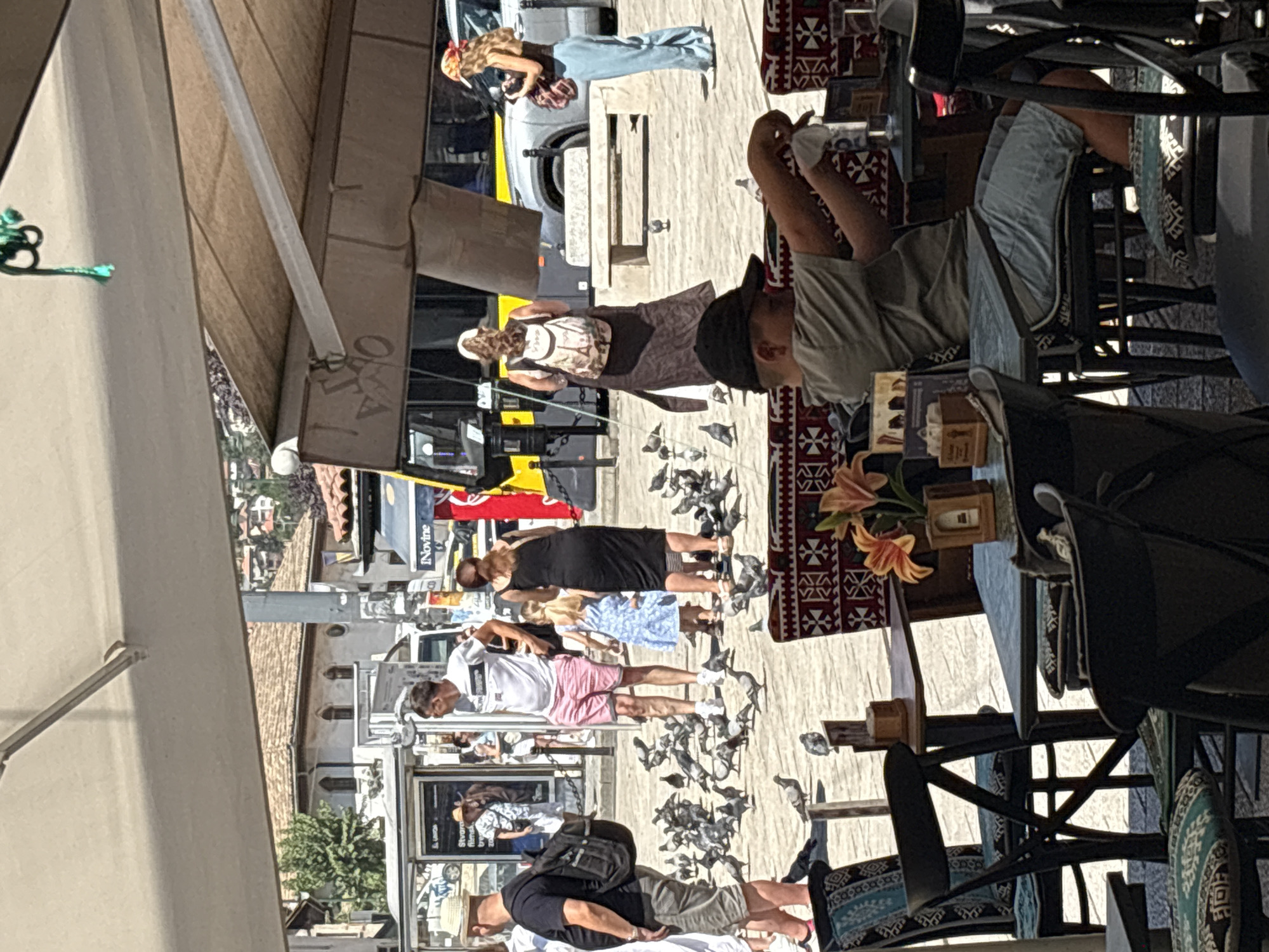 A group of people are gathered in a public square, likely in Sarajevo, Bosnia and Herzegovina, on a sunny day. In the foreground, a young boy with a black cap sits at a table, his arms extended as he interacts with something off-camera. He is wearing a grey t-shirt and light blue shorts. Several empty chairs with patterned cushions are visible around the table.
In the mid-ground, a flock of pigeons occupies the paved ground. Among the pigeons, several people are standing or walking. To the left, a man wearing a straw hat, dark t-shirt, and khaki shorts with a backpack walks away from the camera. Next to him, a man in a white t-shirt and pink shorts stands facing a woman in a black dress, who is holding a young girl in a blue dress. Further to the right, a woman in a dark dress and white headscarf, carrying a backpack, walks away. Another woman with blonde hair and a red bandana, wearing a dark top and light blue trousers, stands holding a bag.
In the background, a yellow and black bus is parked, with buildings and rooftops visible beyond. An awning covers part of the square, providing shade. The overall scene depicts a vibrant public space with people enjoying the outdoors, with pigeons as a common presence.