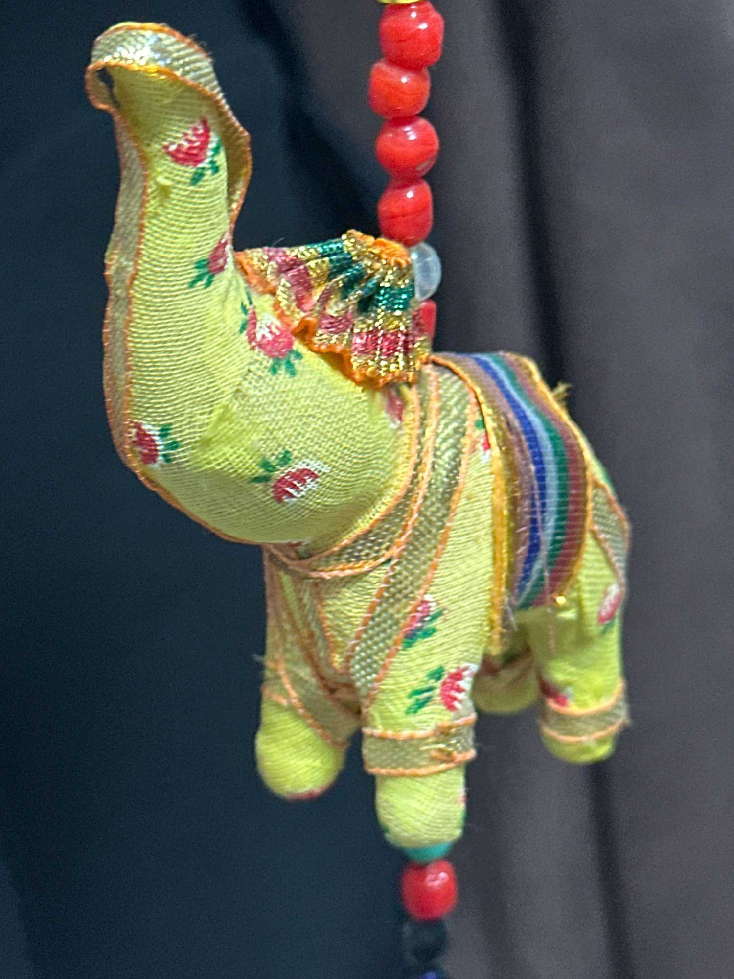 A close-up, eye-level shot showcases a small, fabric elephant ornament hanging against a dark, out-of-focus background. The elephant is predominantly lime green with a pattern of small red roses. It is adorned with colorful, striped fabric draped over its back and has metallic gold trim along its edges. The elephant is suspended by a string of red beads, with additional beads visible above and below the ornament. The image is devoid of any specific geographical markers linking it to Nevinnomyssk, Russia.