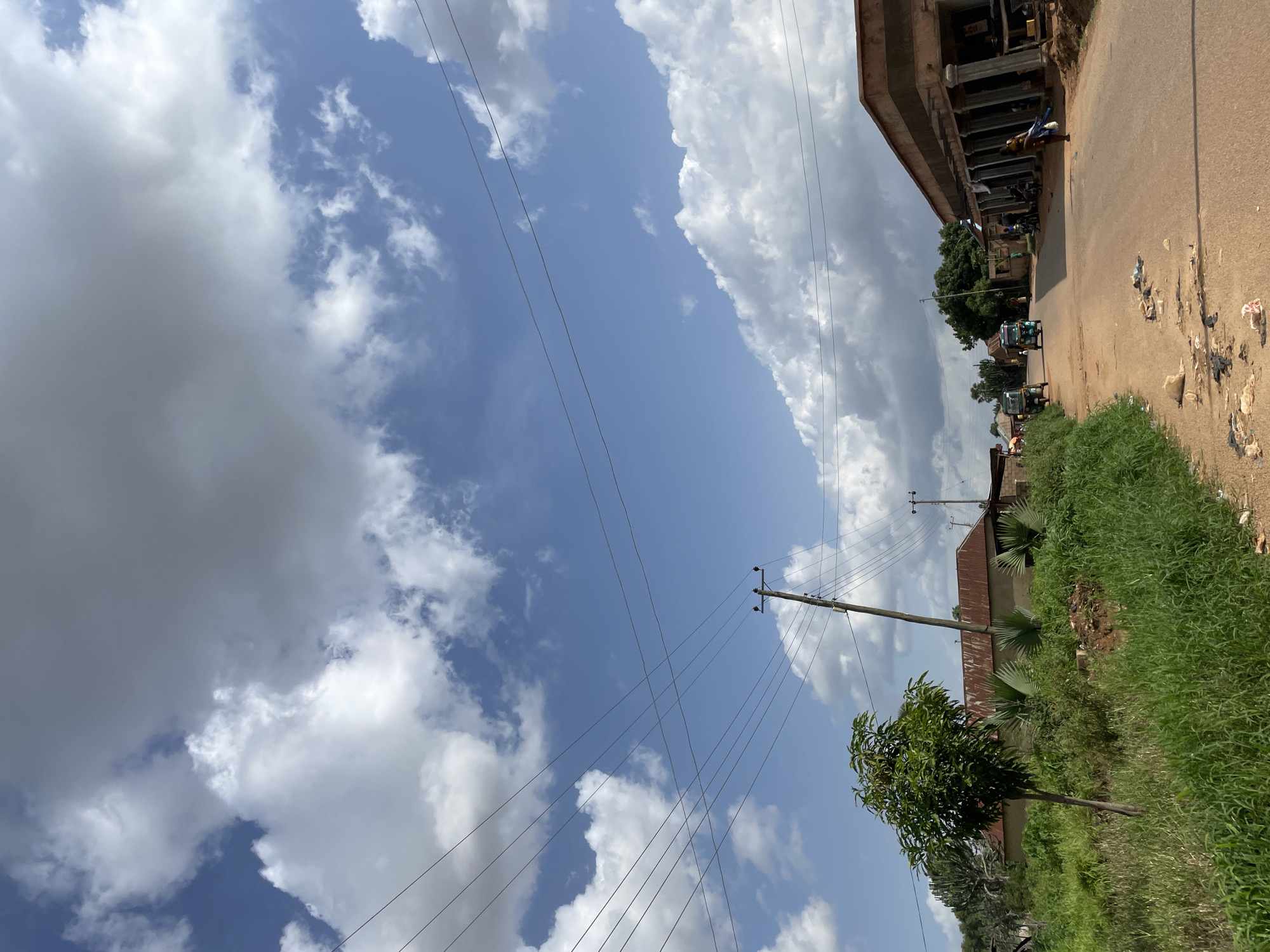 The image captures a street scene in Udu, Nigeria, under a partly cloudy sky. The road is unpaved and appears dusty, with scattered litter along its edges. On the left side of the road, there is overgrown green grass and a small tree. Further back, several buildings with corrugated iron roofs are visible, along with utility poles and electrical wires crisscrossing the sky. On the right side, a building under construction or with an unfinished facade features a row of columns. Several people are present in the scene; one individual is walking on the right side of the road, carrying items, and two individuals are seen near the buildings. A few small vehicles, possibly auto-rickshaws, are present on the road. The overall environment suggests a semi-urban or rural setting within Udu.
