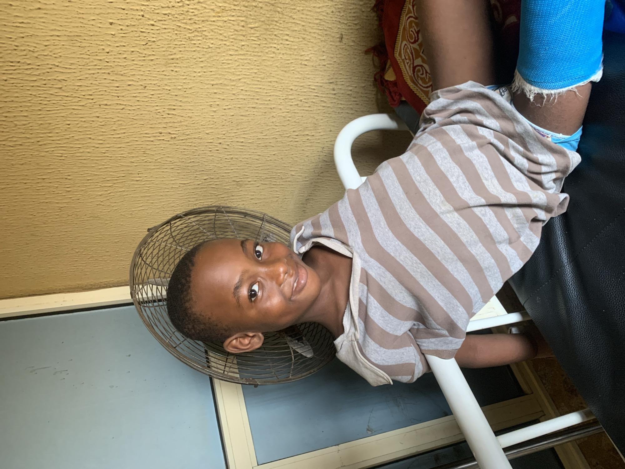 The image shows a young Black child lying down, with their head resting on a fan. The child is wearing a striped shirt and has a blue cast on their leg. The background appears to be a room with a textured yellow wall and a black leather couch. A colorful patterned cloth is visible on the couch. The lighting suggests it is daytime, and there are no clear weather indicators. The child is looking directly at the camera with a slight smile, conveying a sense of comfort or playfulness despite the cast. The presence of the fan and the informal setting suggest a domestic environment in Shagamu, Nigeria.