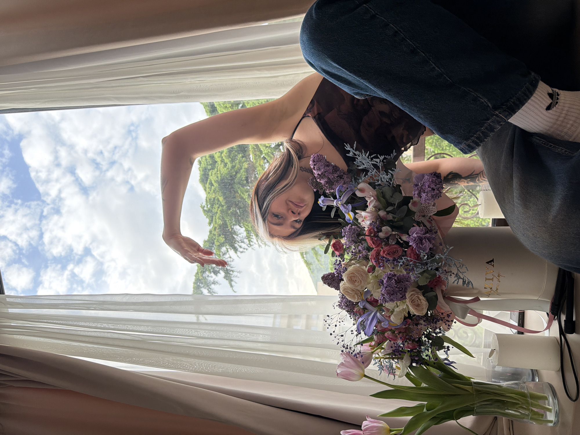 The image captures a young woman posing indoors by a large window with a view of a bright, cloudy sky and lush green trees, characteristic of a scenic location like Budva, Montenegro. The photograph is oriented vertically, showing the subject and scene from a low angle.
The woman, with multi-toned brunette hair featuring lighter streaks, is seen from the waist up. She wears a dark, patterned sleeveless top and blue jeans. A nose piercing and a tattoo on her right forearm are visible. She looks directly at the camera with a slight, pleasant expression, while her left hand is raised and open, almost reaching out.
A prominent feature is a large, elaborate bouquet of various flowers, including roses, irises, lilacs, and other blossoms in shades of purple, pink, white, and cream. This bouquet is arranged in a tall, cylindrical white box, decorated with a pink ribbon. The text "VIVA flower shop" is visible on the side of the box. Nearby, a glass vase holds several light pink tulips with green stems. Two white cylindrical candles are also on the surface next to the flower arrangements.
Through the window, a bright daytime sky with soft white clouds can be seen. Tall green trees dominate the outdoor view, suggesting a natural and verdant environment. The indoor setting features white sheer curtains, and heavier beige-brown curtains pulled to the sides. The woman's white socks, adorned with a black Adidas trefoil logo, are also visible. The overall mood is bright and natural, indicating a clear, possibly slightly overcast, day.