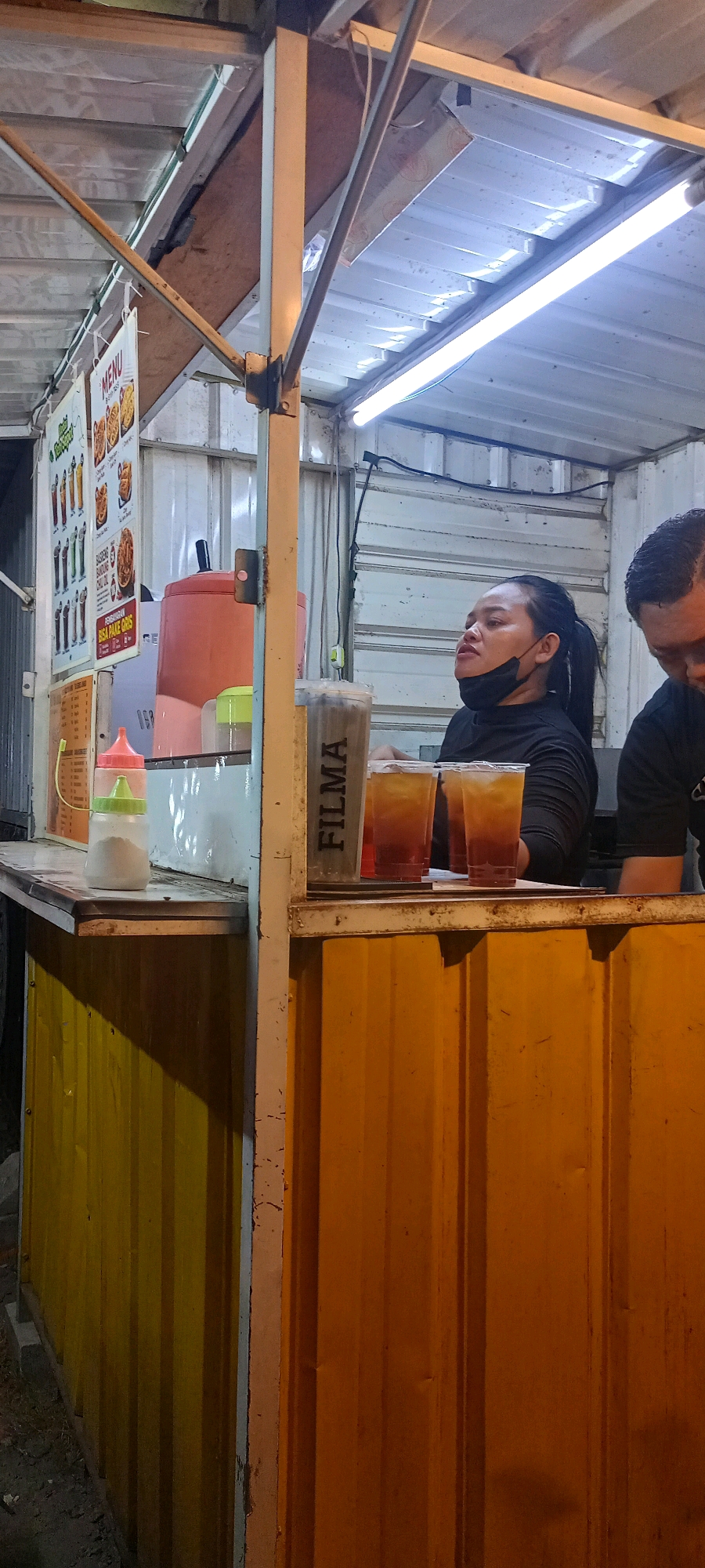 An outdoor food stall in Bendotretek, Indonesia, is depicted at night. The stall features a corrugated yellow and orange metal exterior. Inside, a woman with a black ponytail and a black face mask pulled down to her chin, wearing a black long-sleeved shirt, is visible. She is looking upwards and to the left. Next to her, a man in a black shirt is partially visible on the right side of the frame, with his head bowed. On the counter, there are three plastic cups of iced tea, one of which has the word "FILMA" printed vertically on its side. A pink dispenser and a white dispenser with a green cap are also on the counter, along with a menu board displaying images of food items. Fluorescent lights illuminate the scene from the ceiling.